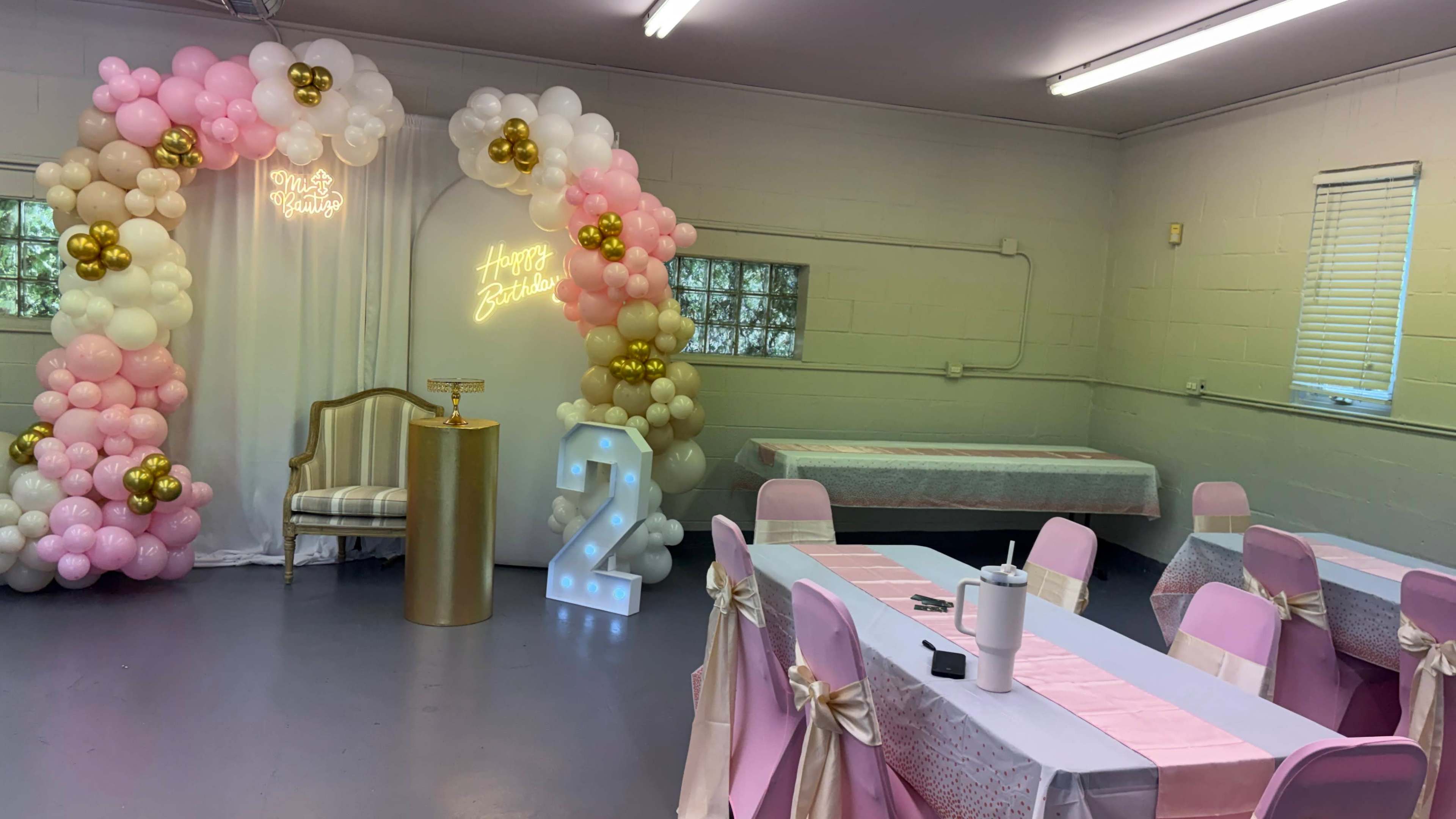The image shows a decorated room for a birthday celebration, featuring a balloon arch, a golden table, and a large "2" illuminated sign.