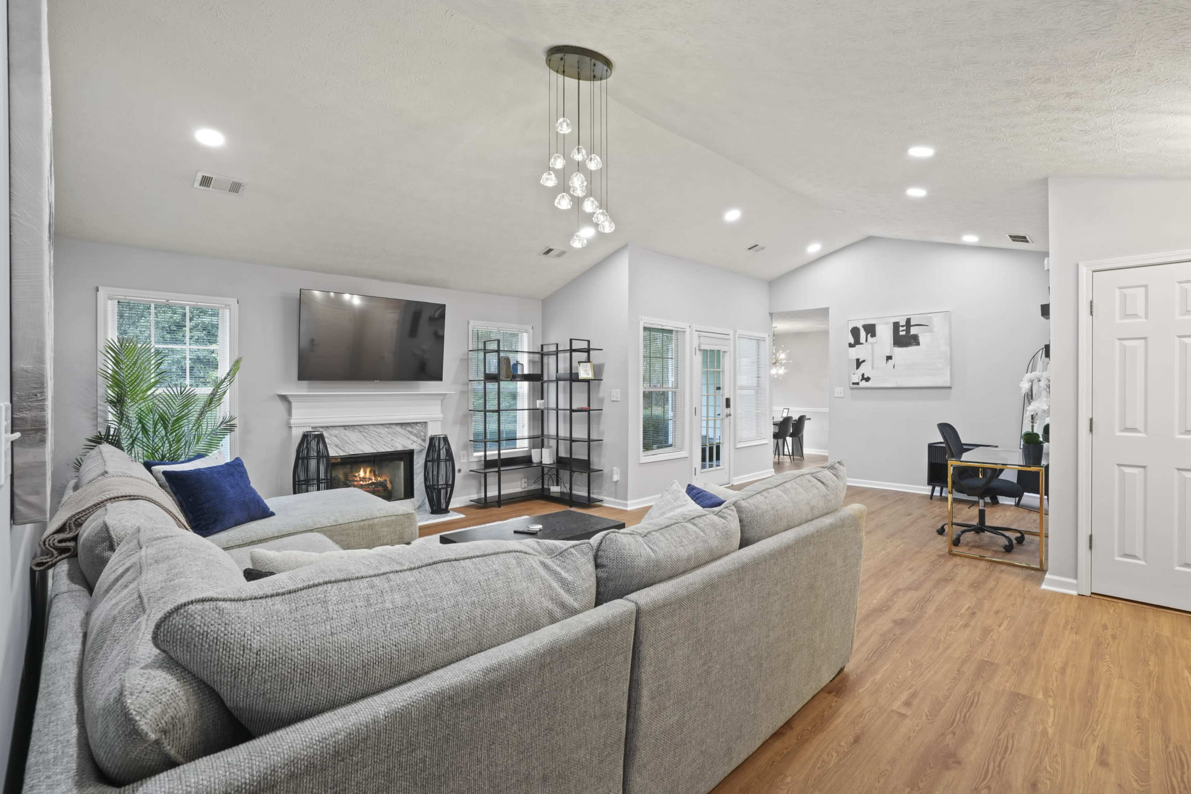 The image shows a modern living room with a gray sectional sofa, a fireplace, a television mounted on the wall, and a small workspace with a desk and chair.