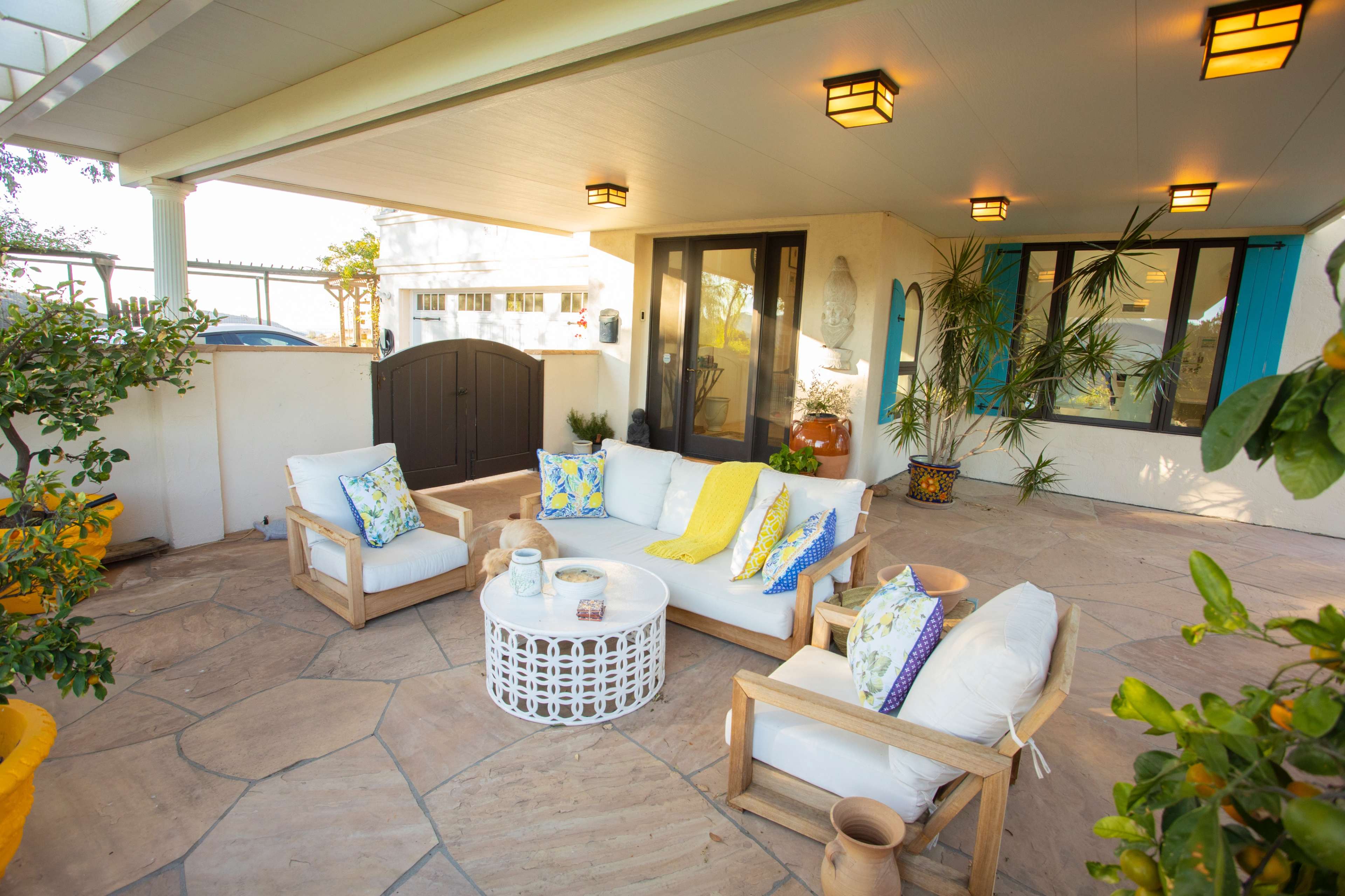 The image shows a cozy outdoor seating area with a white couch and wooden chairs, arranged around a round coffee table on a stone patio.