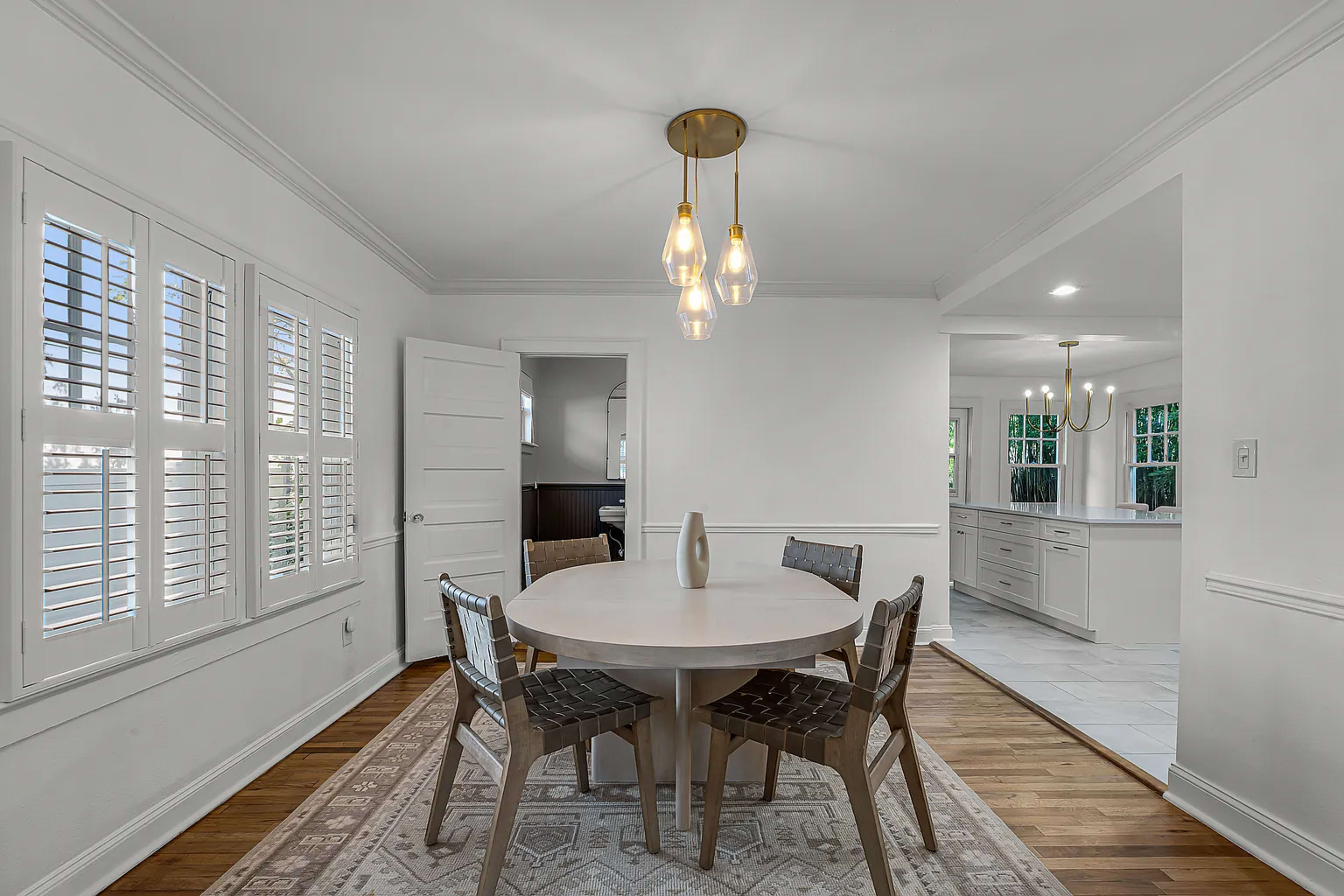 The image shows a dining room with a round table, four chairs, and a light fixture, featuring a doorway leading to another room and large windows with shutters.