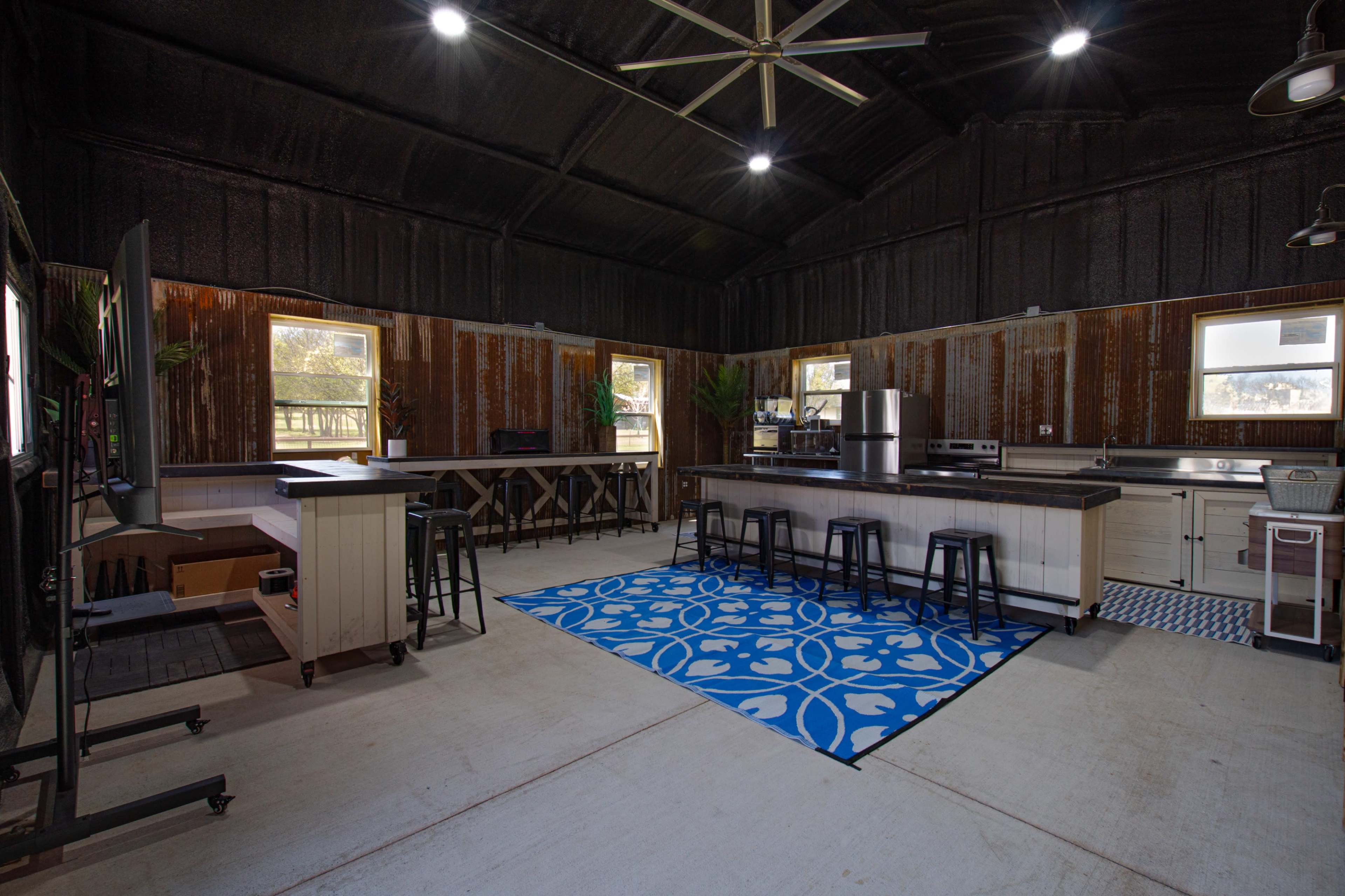 The image shows a modern kitchen inside a rustic building with corrugated metal walls, featuring a wide island, bar stools, and blue patterned rugs.