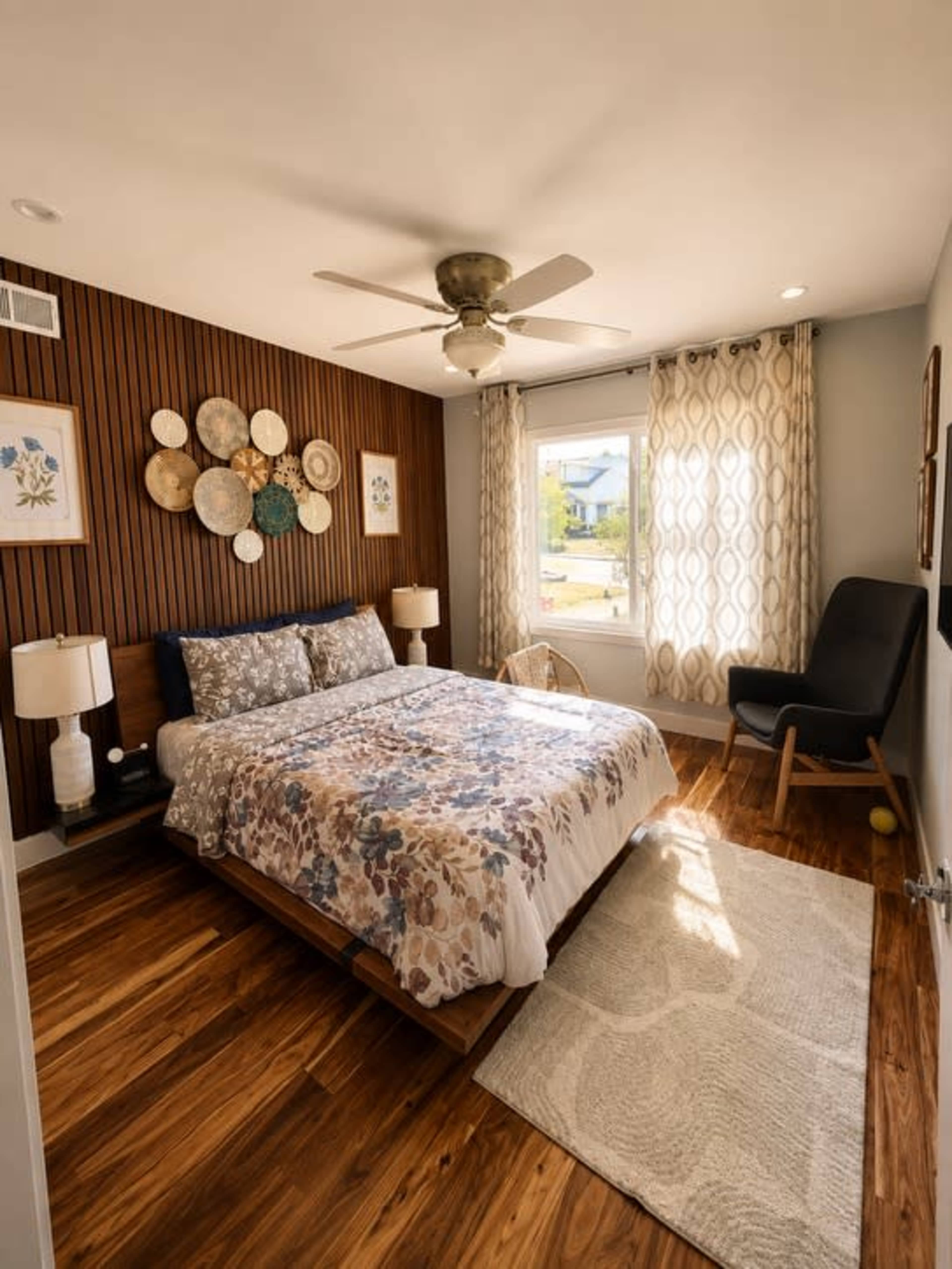 The image shows a well-lit bedroom featuring a bed with a floral-patterned duvet, wooden wall accents, and artwork arranged above the bed.