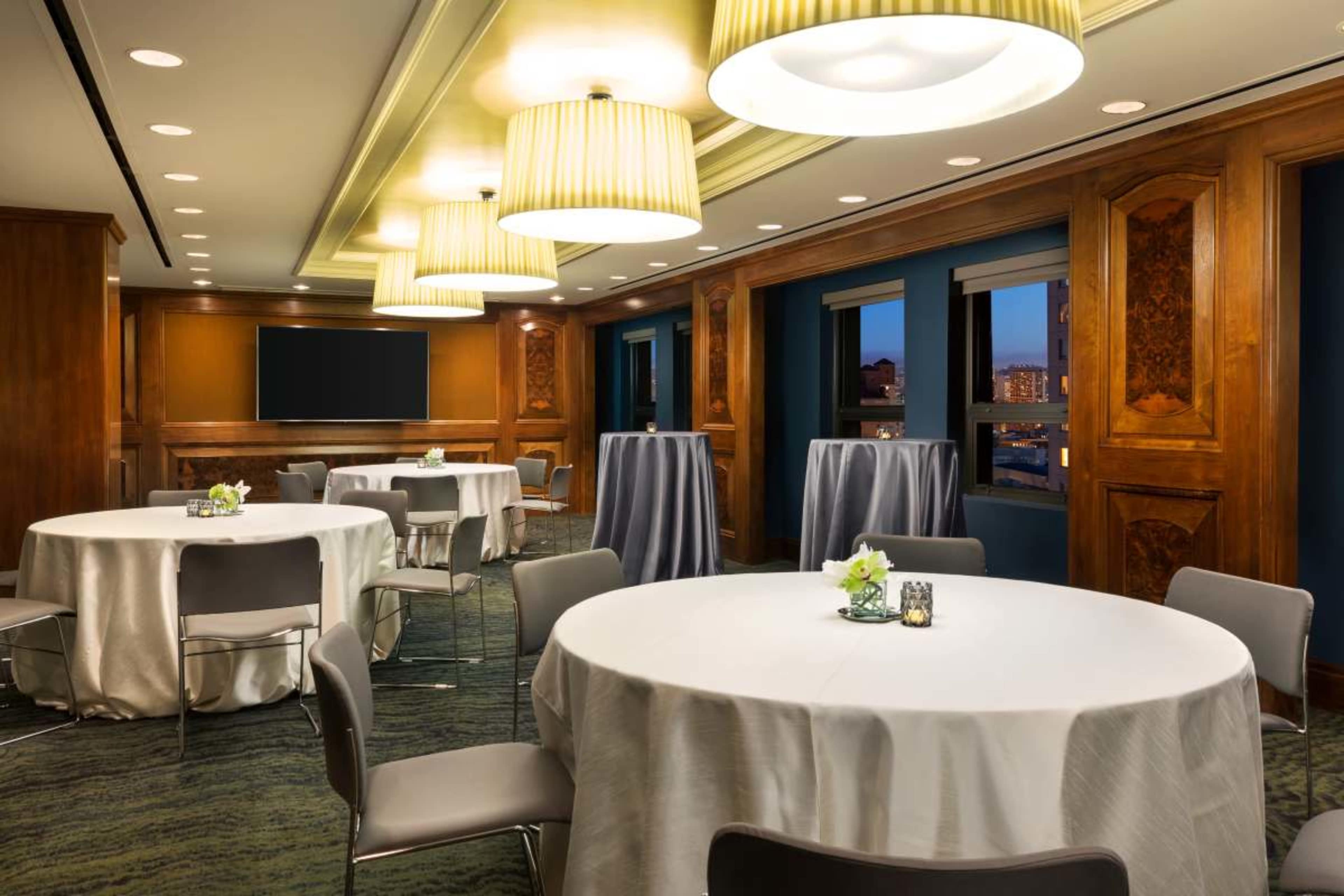 The image shows a well-lit event space featuring round tables with light-colored tablecloths and tall cocktail tables, surrounded by elegant decor and windows overlooking an urban skyline.