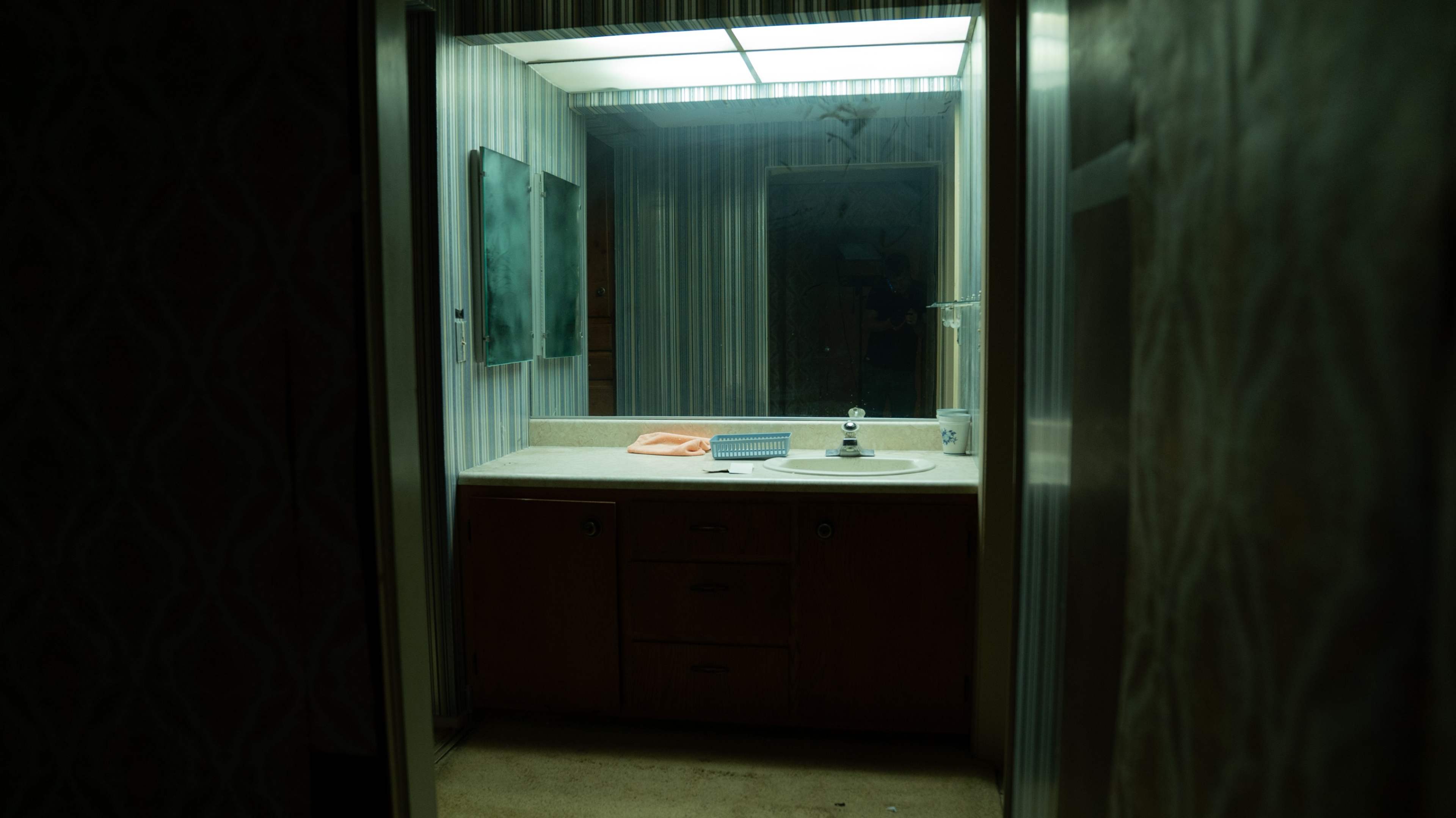 The image shows a dimly lit bathroom with a sink, a countertop, and a large mirror surrounded by striped wallpaper.