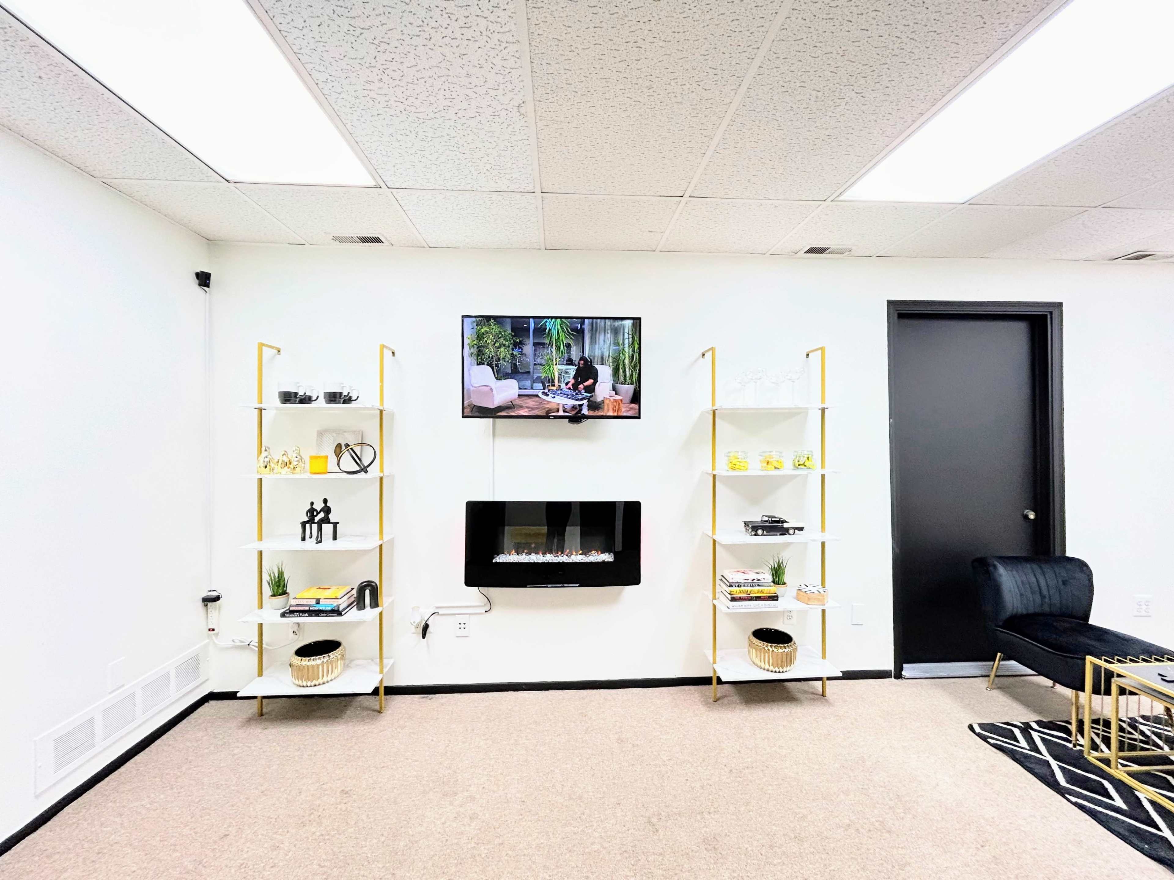 A minimalist room with a mounted TV above a wall-mounted electric fireplace, flanked by two white shelving units, and a black armchair on a patterned rug.