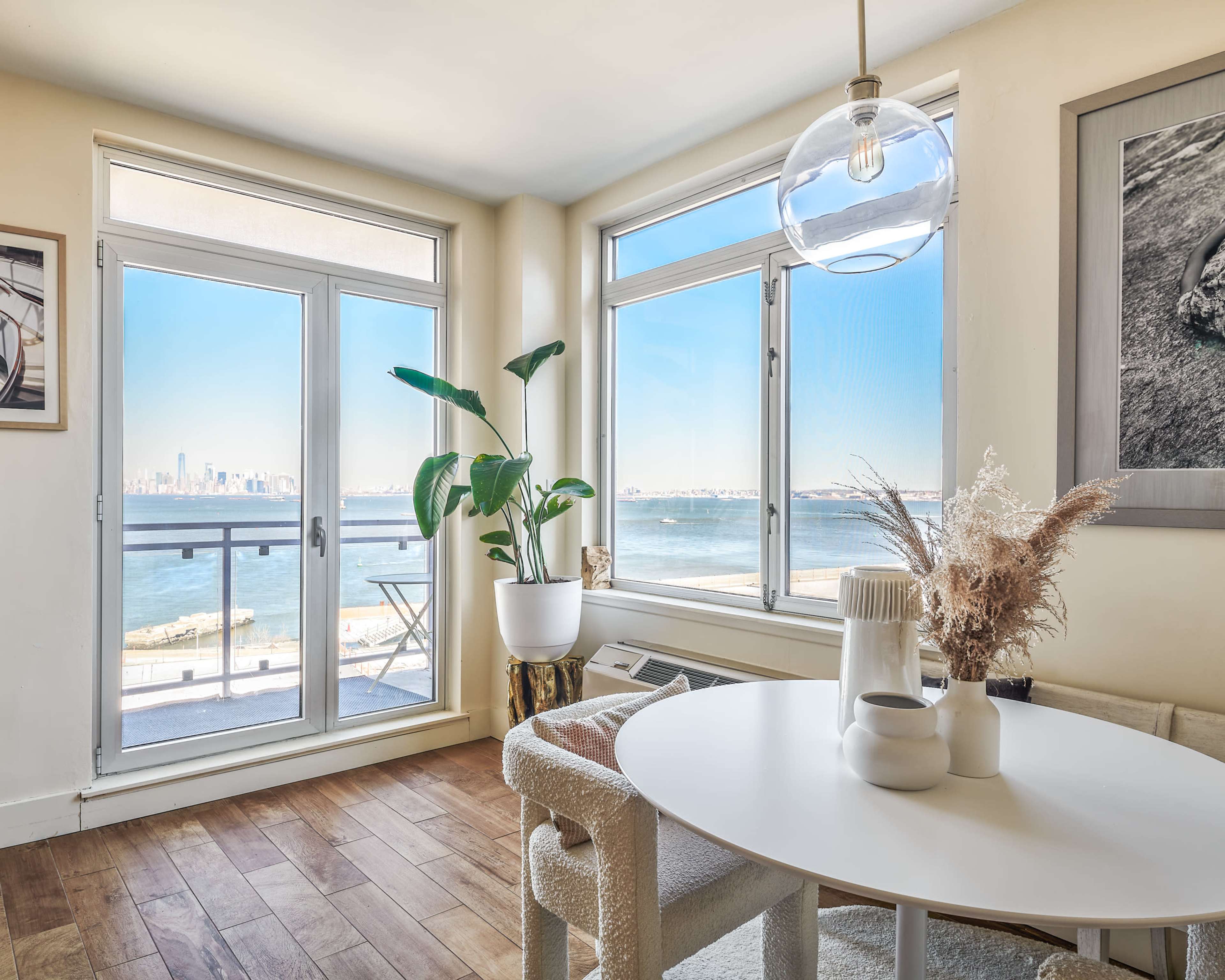 A cozy living area with a round table, potted plant, and large windows showcasing a view of the water and city skyline.