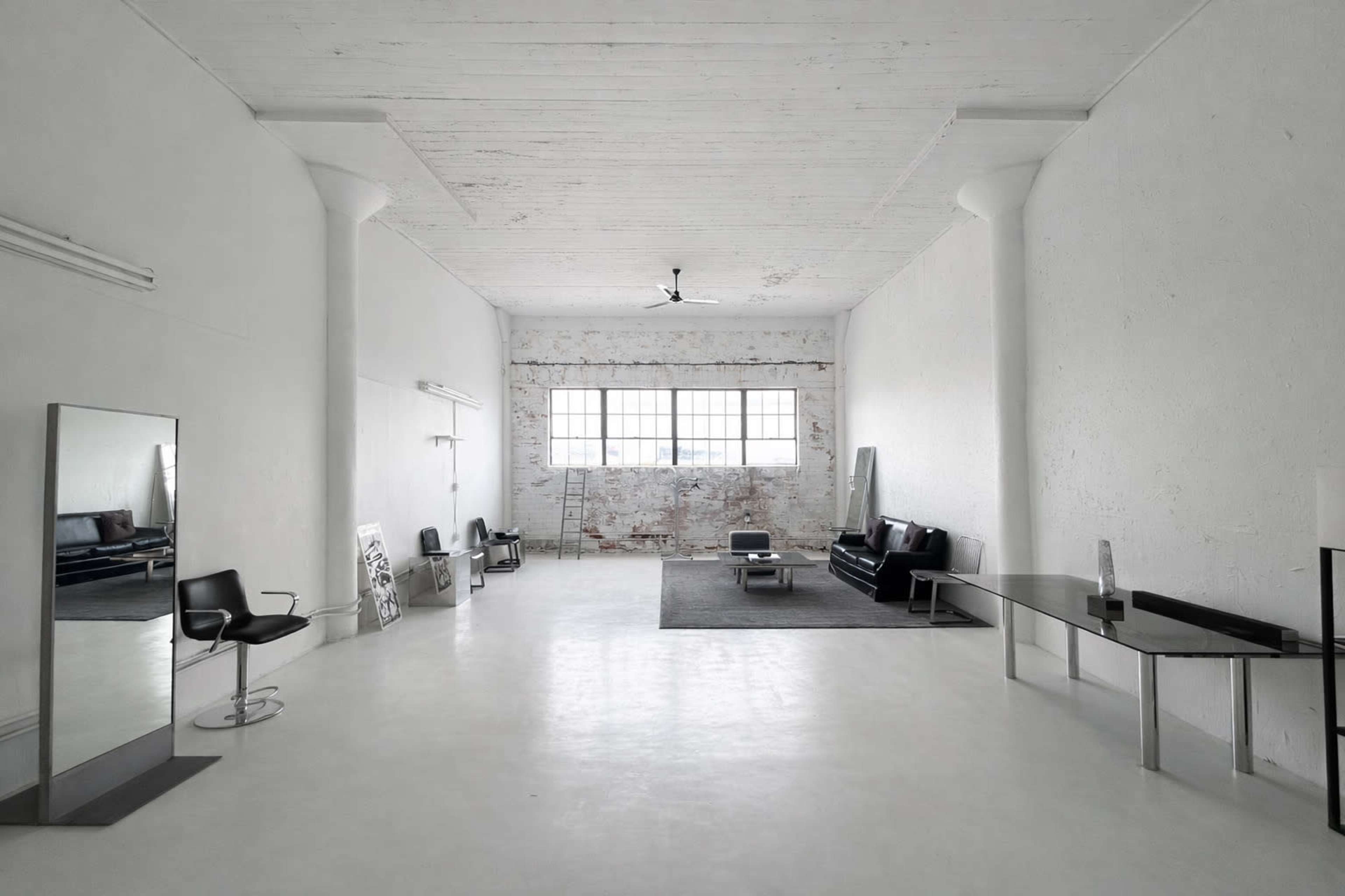 The image shows a spacious, minimalist interior with white walls, large windows, and modern furniture including black sofas and a sleek table.