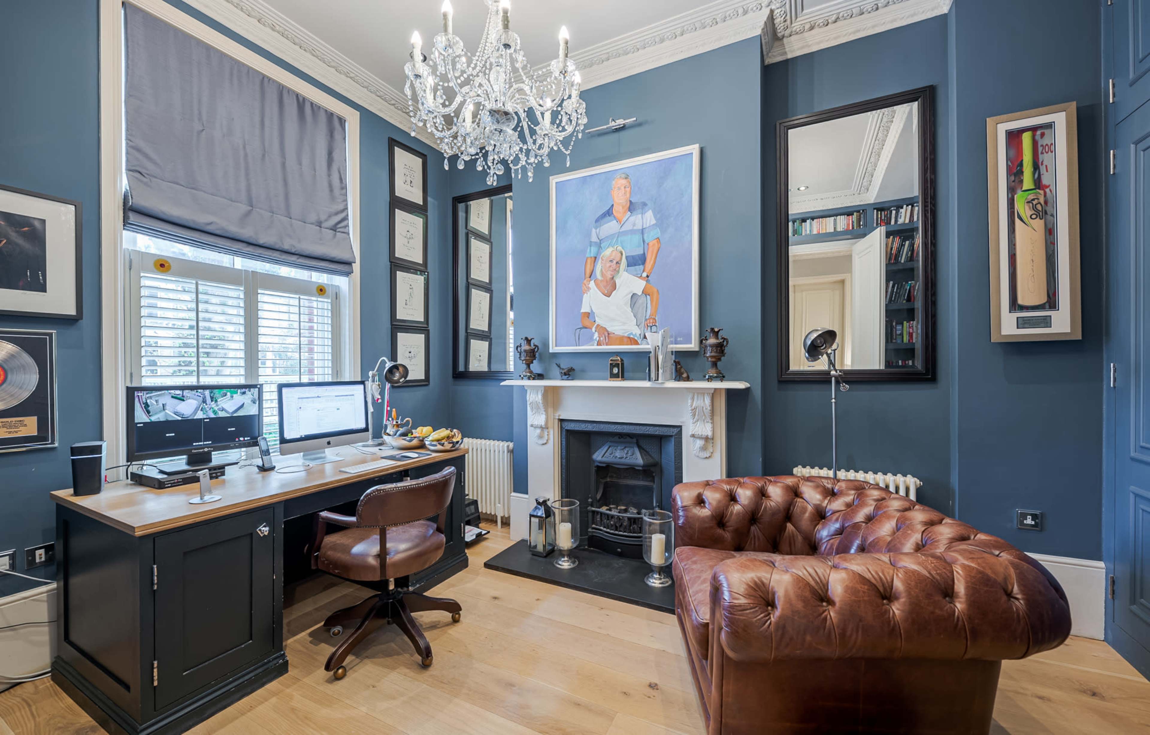 The image shows a well-decorated home office featuring a wooden desk with a computer, a leather armchair, and a fireplace surrounded by framed art and awards.