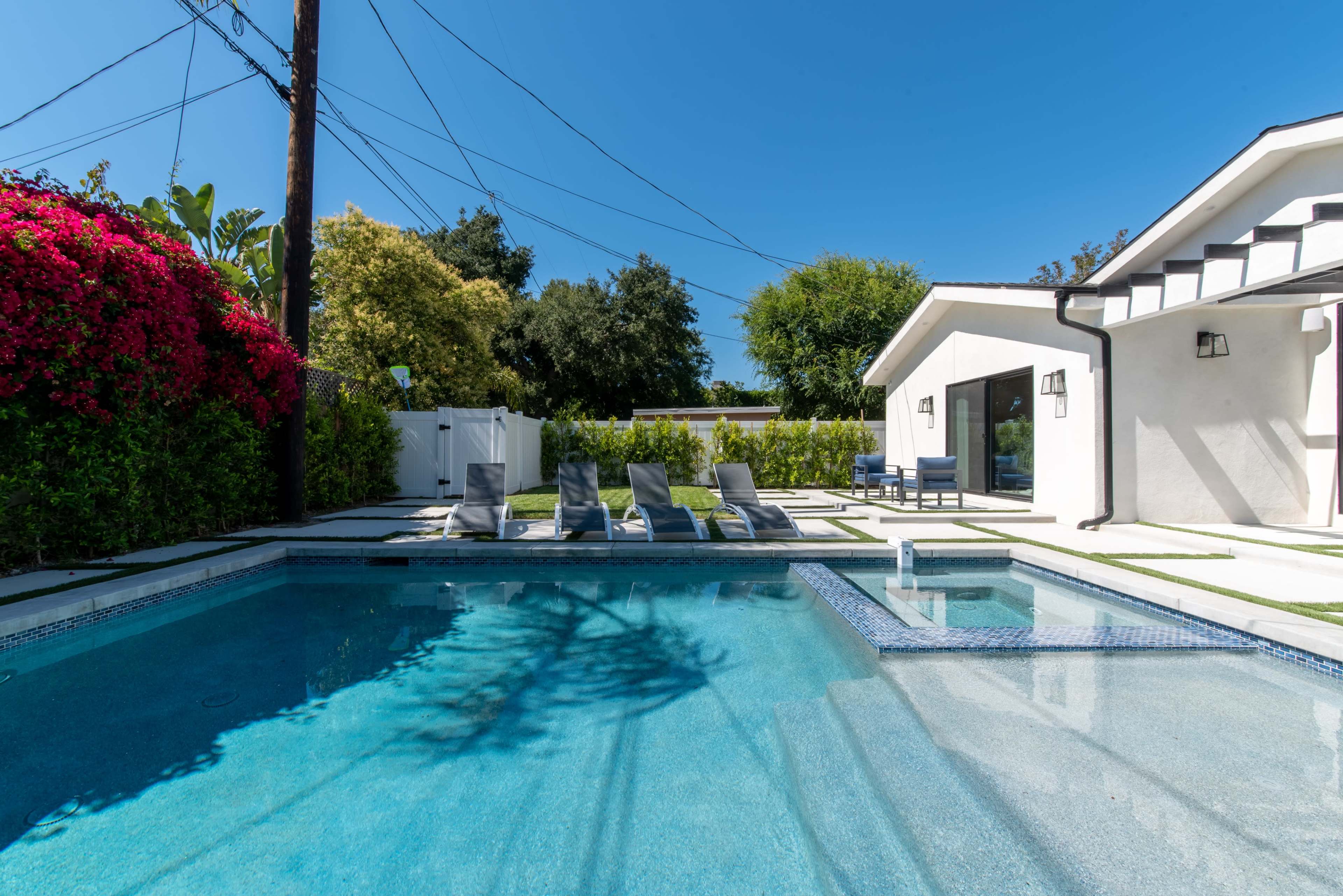 The image shows a swimming pool surrounded by lounge chairs and vibrant greenery, with a house and power lines in the background.