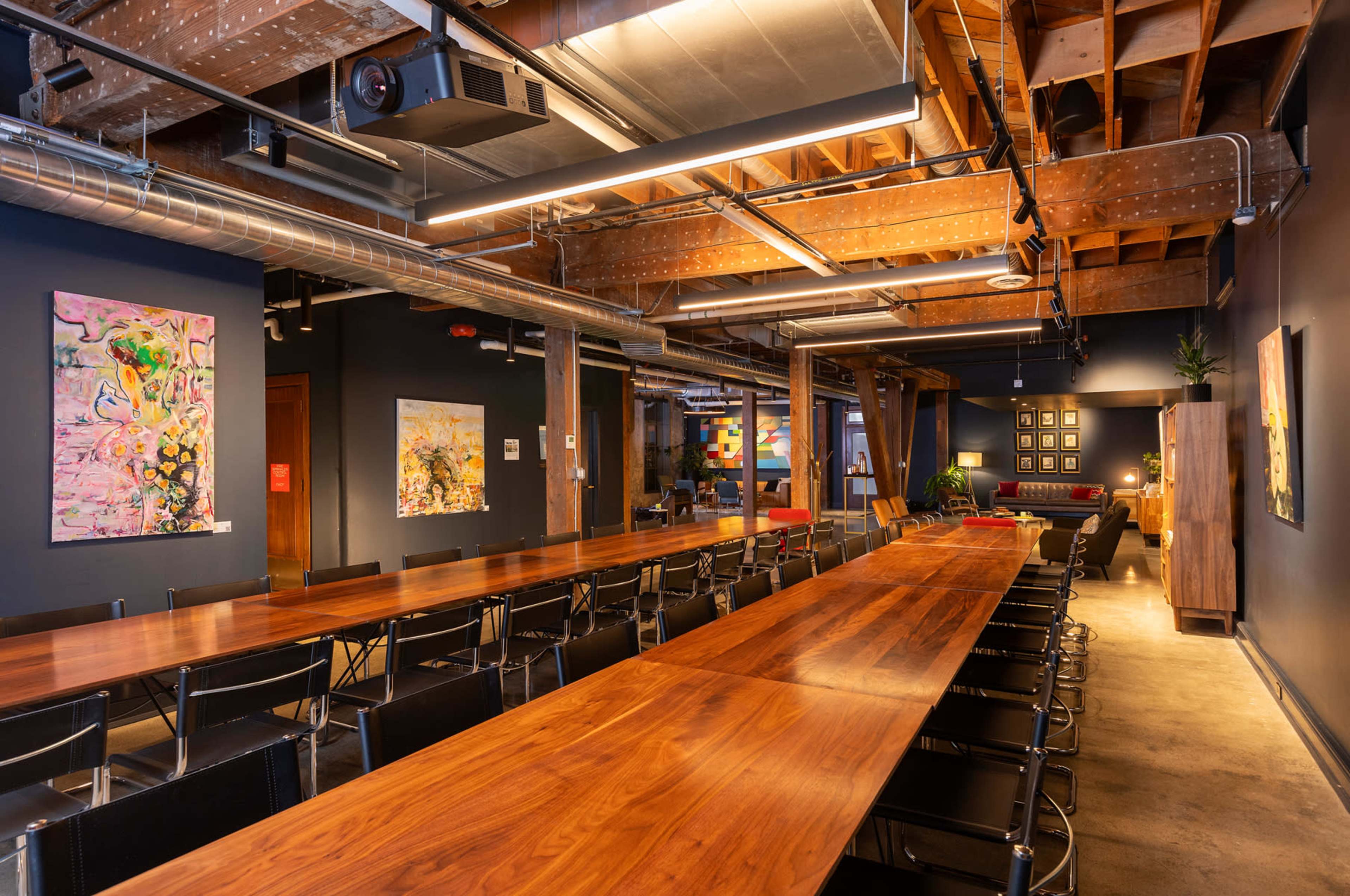 The image shows a spacious interior of a modern event space with a long wooden table, black chairs, and colorful artwork on the walls.