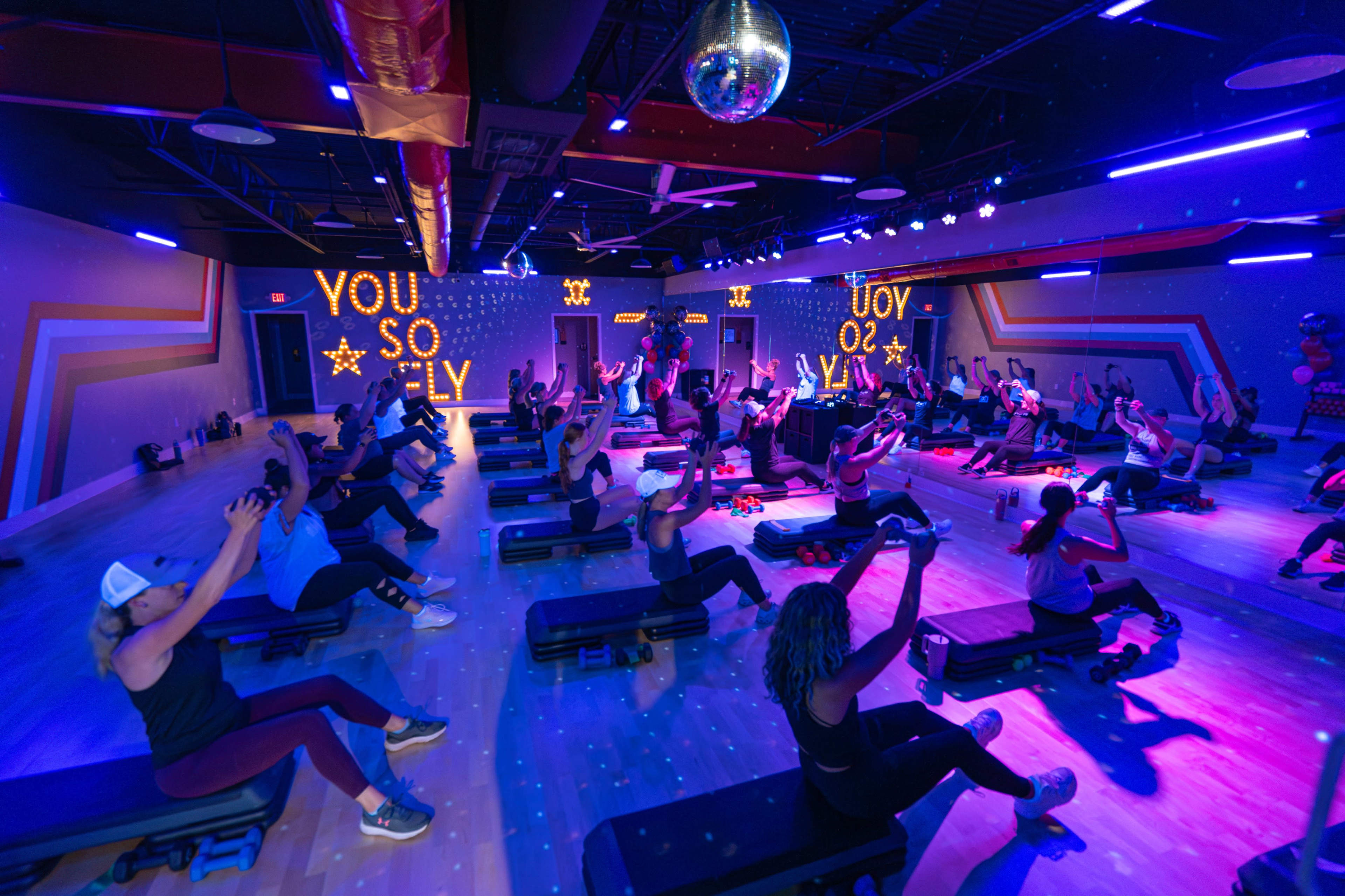 A group of people participates in a fitness class in a brightly lit studio with a disco ball and motivational signs on the walls.