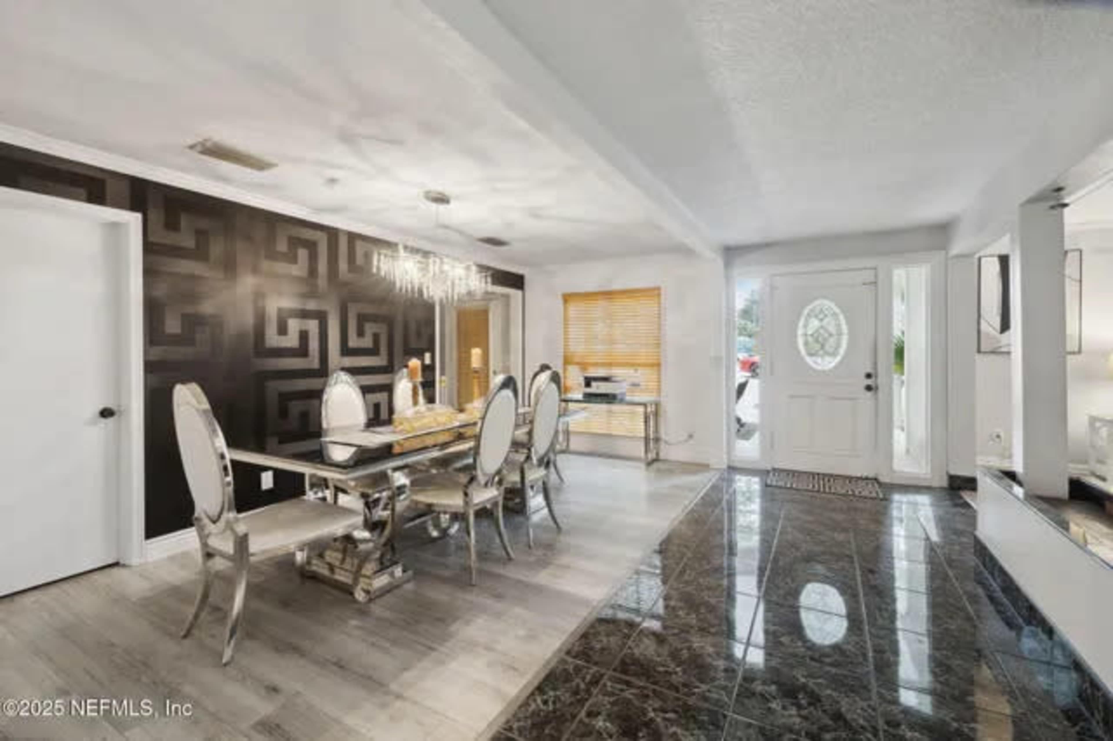 The image shows a modern dining area with a glass dining table and mirrored chairs, adjacent to an entrance featuring a decorative door and large windows.