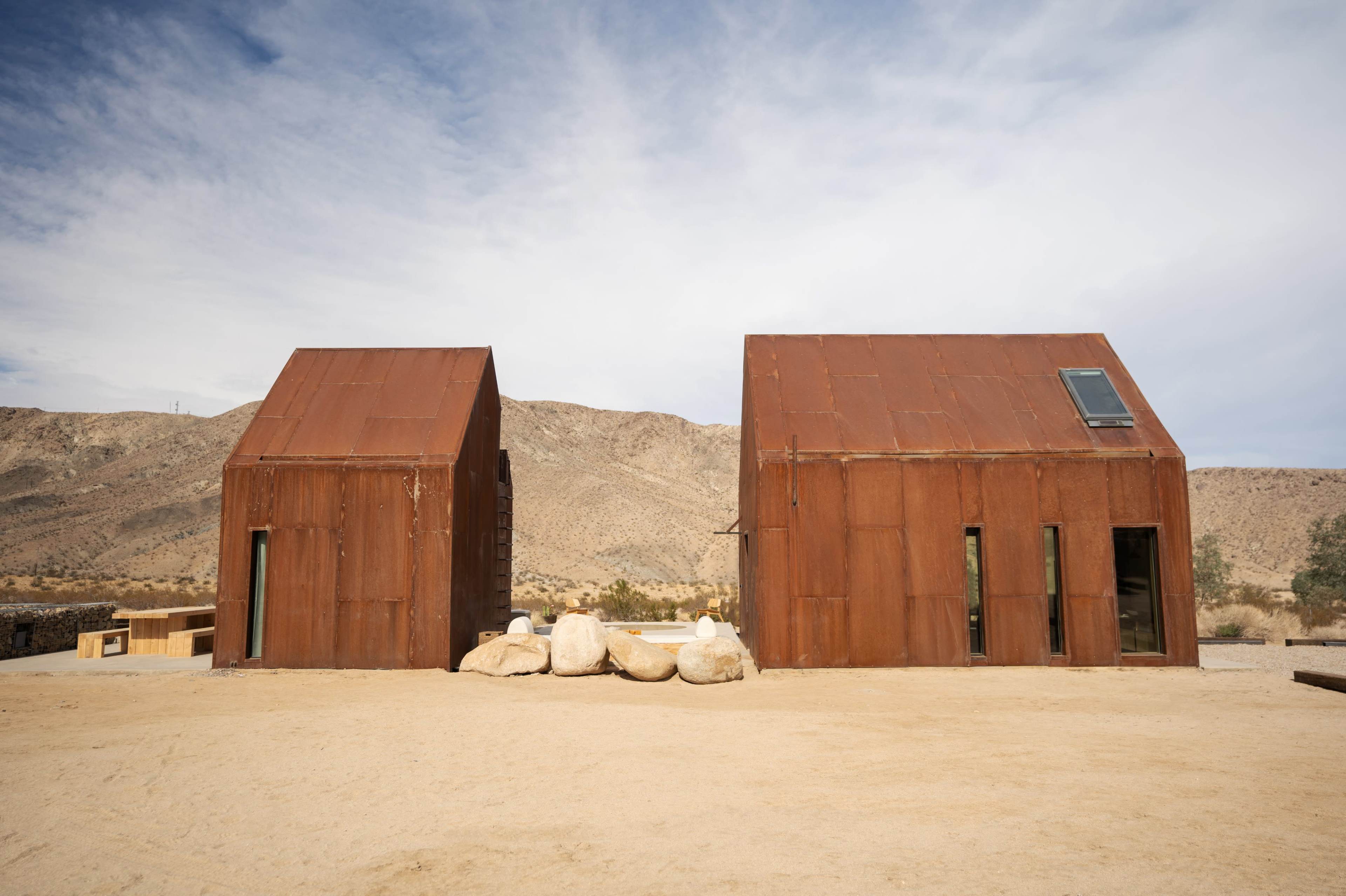 Two rust-colored metal houses are situated in a desert landscape, with rocky terrain and a mountain backdrop.
