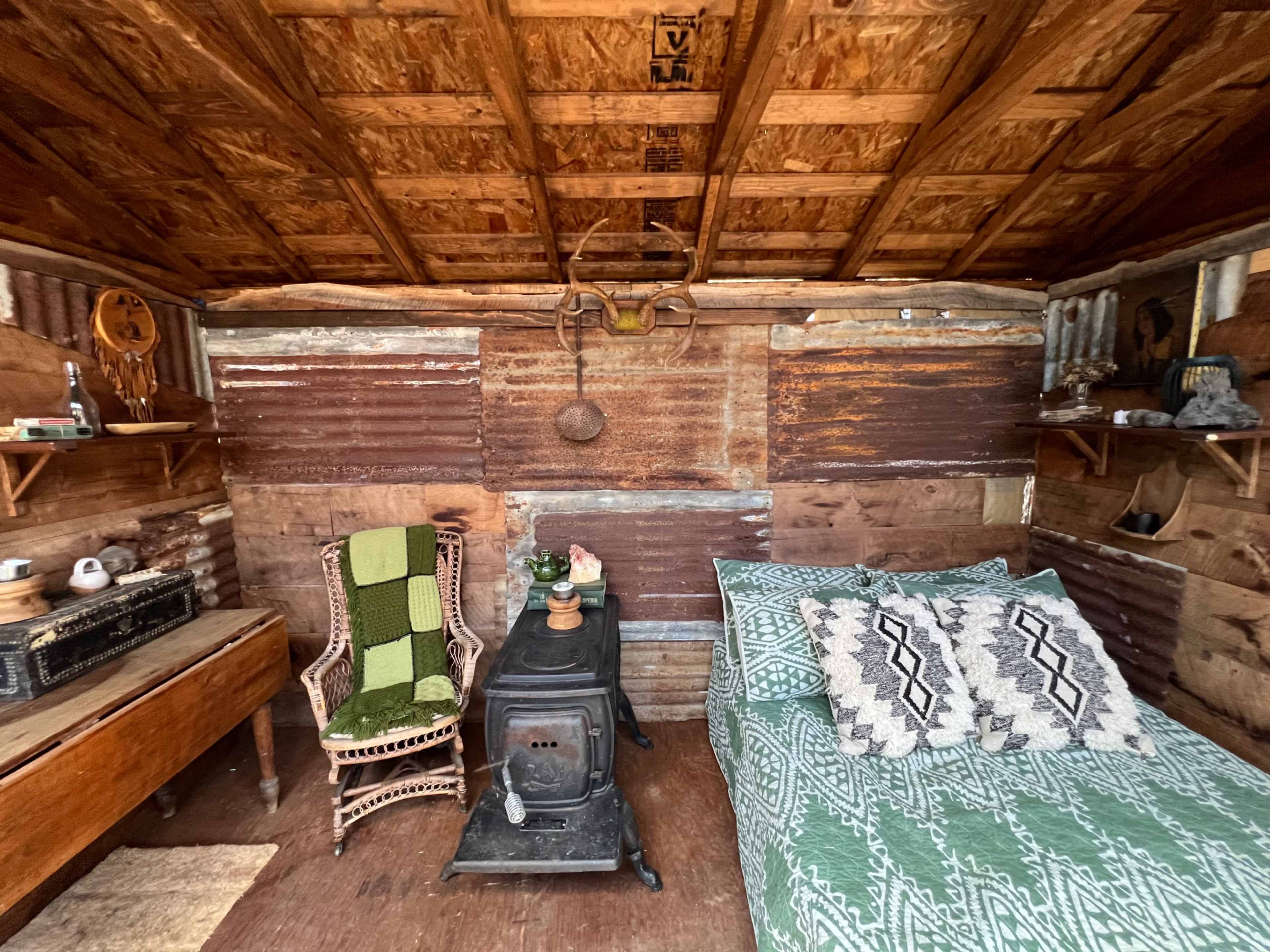 The image shows a rustic interior of a small cabin featuring a bed with a patterned blanket, a wood stove, a wooden table, and shelves against the walls.