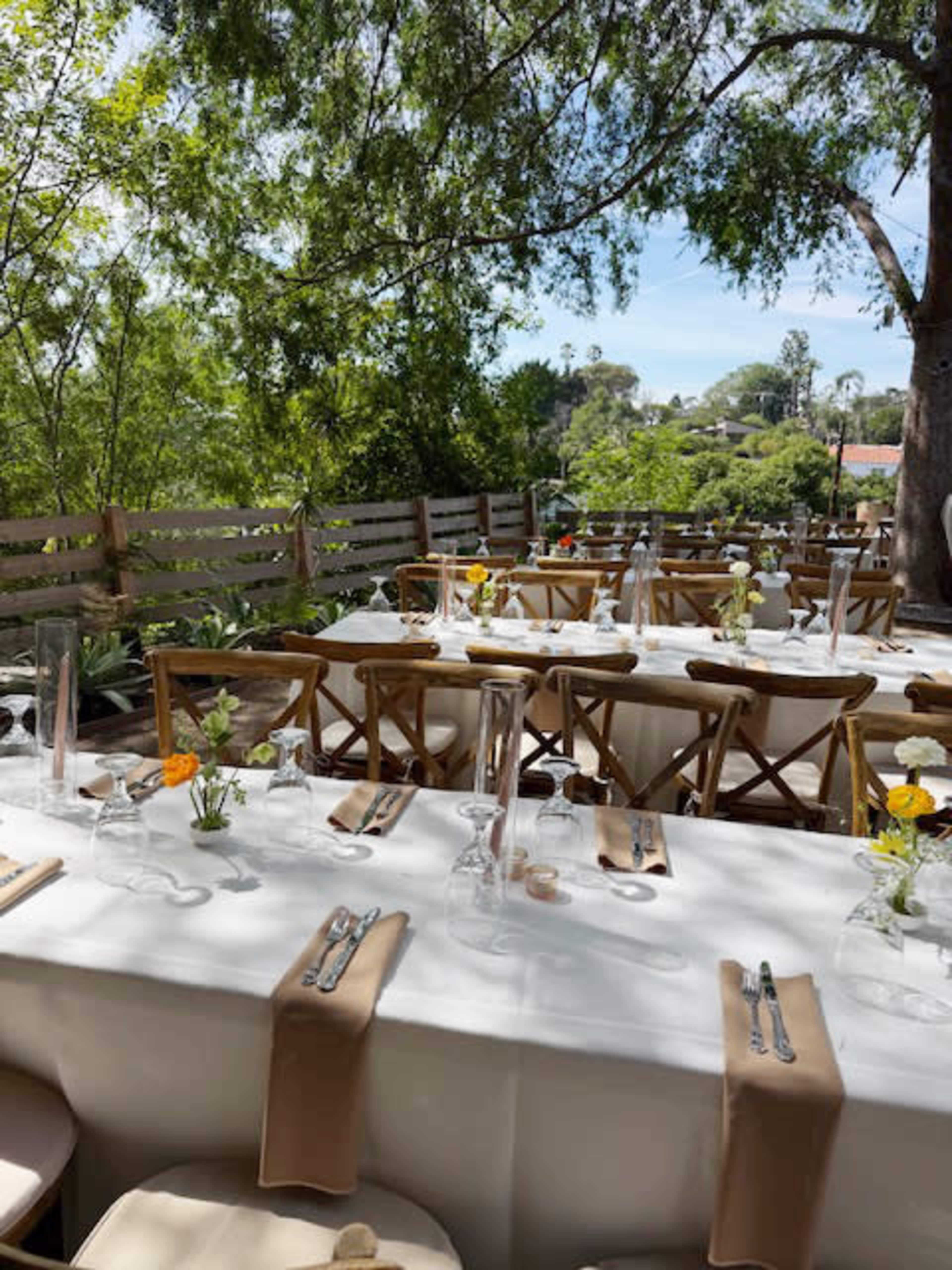 A row of elegantly arranged outdoor tables is set for a dining event, decorated with small flower vases and surrounded by greenery.