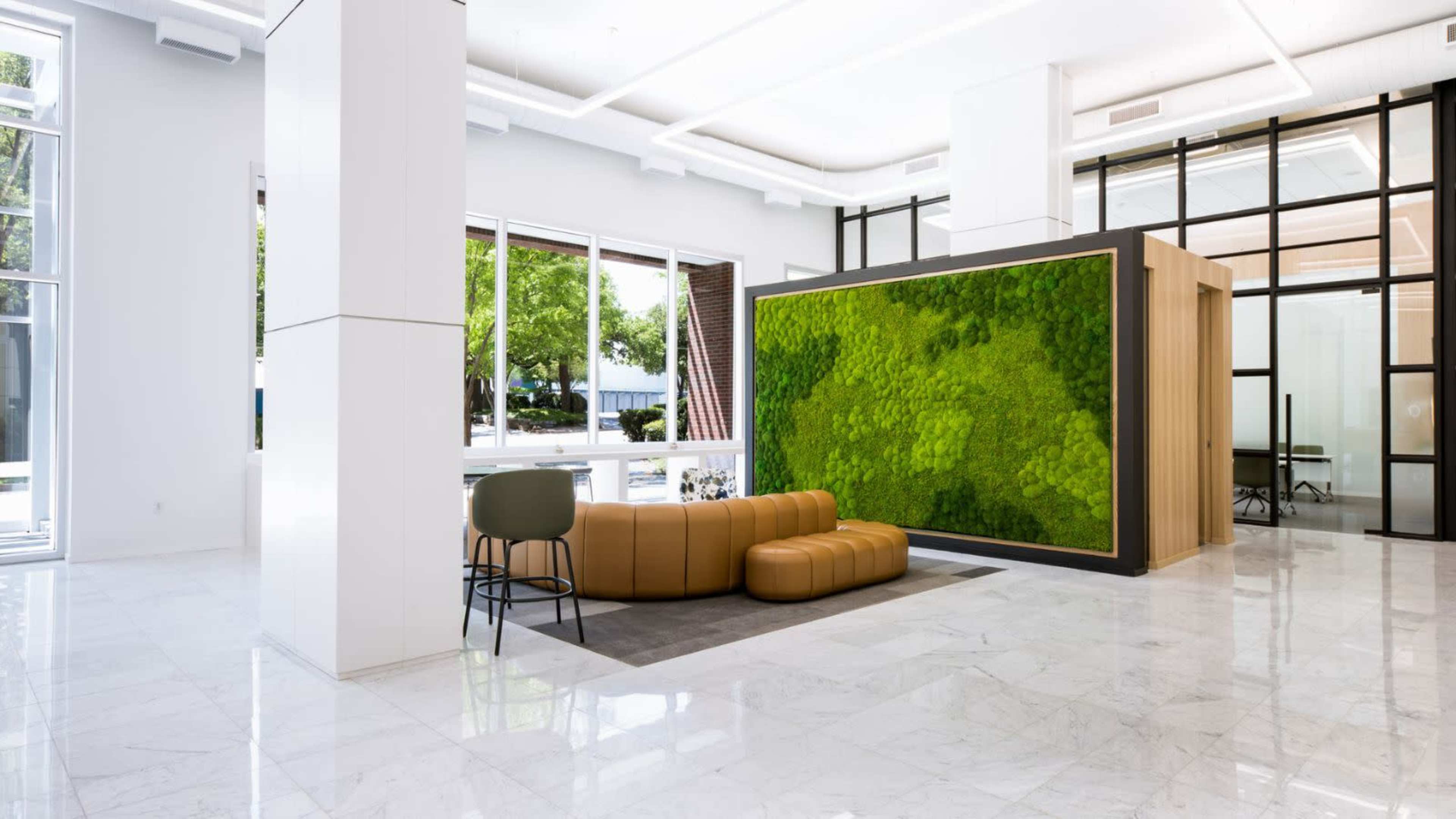The image shows a modern lobby with large windows, a green wall installation, and a comfortable seating area.