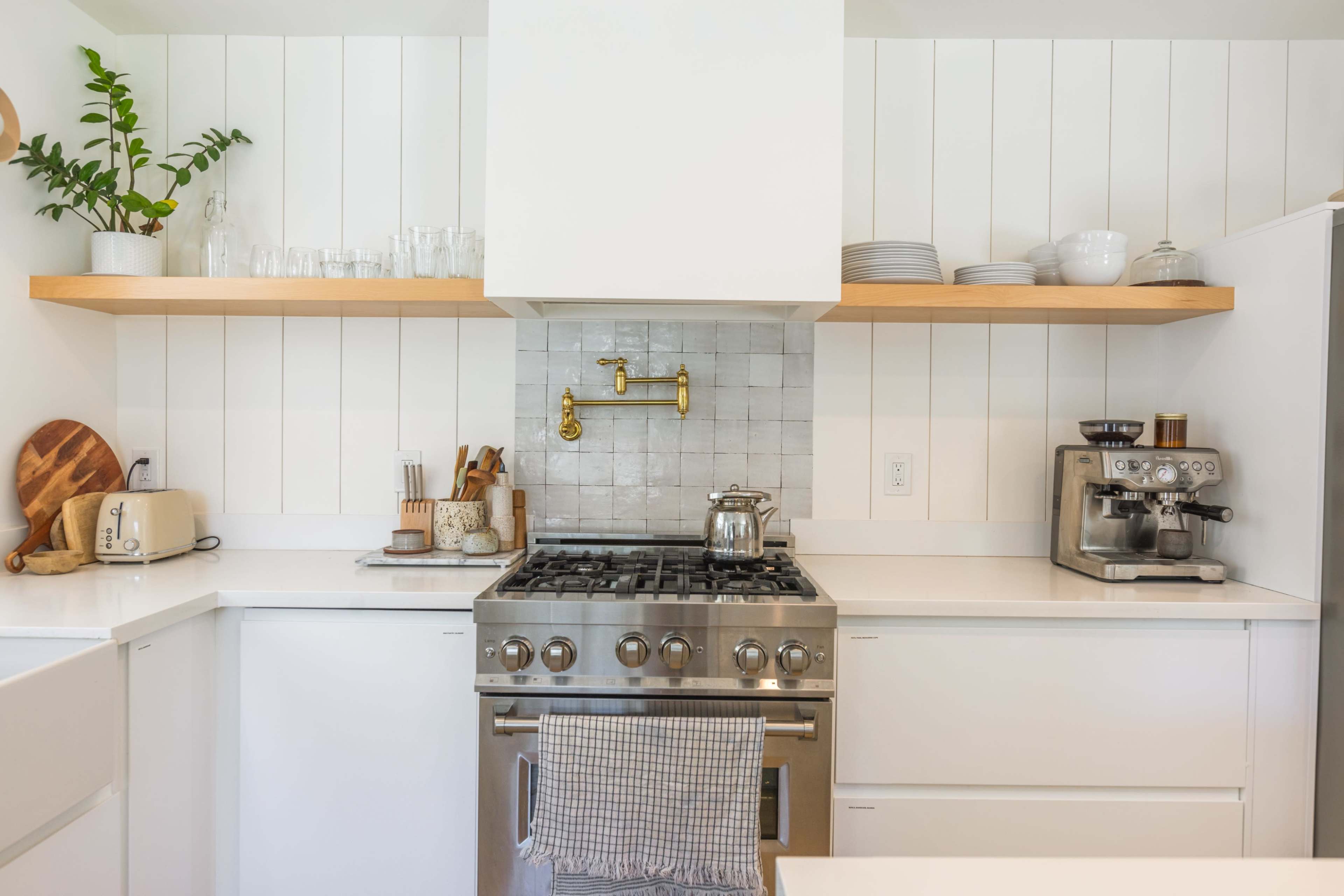A modern kitchen features a gas stove with a stainless steel finish, white cabinetry, wooden shelves holding dishes, and a countertop with a coffee machine.