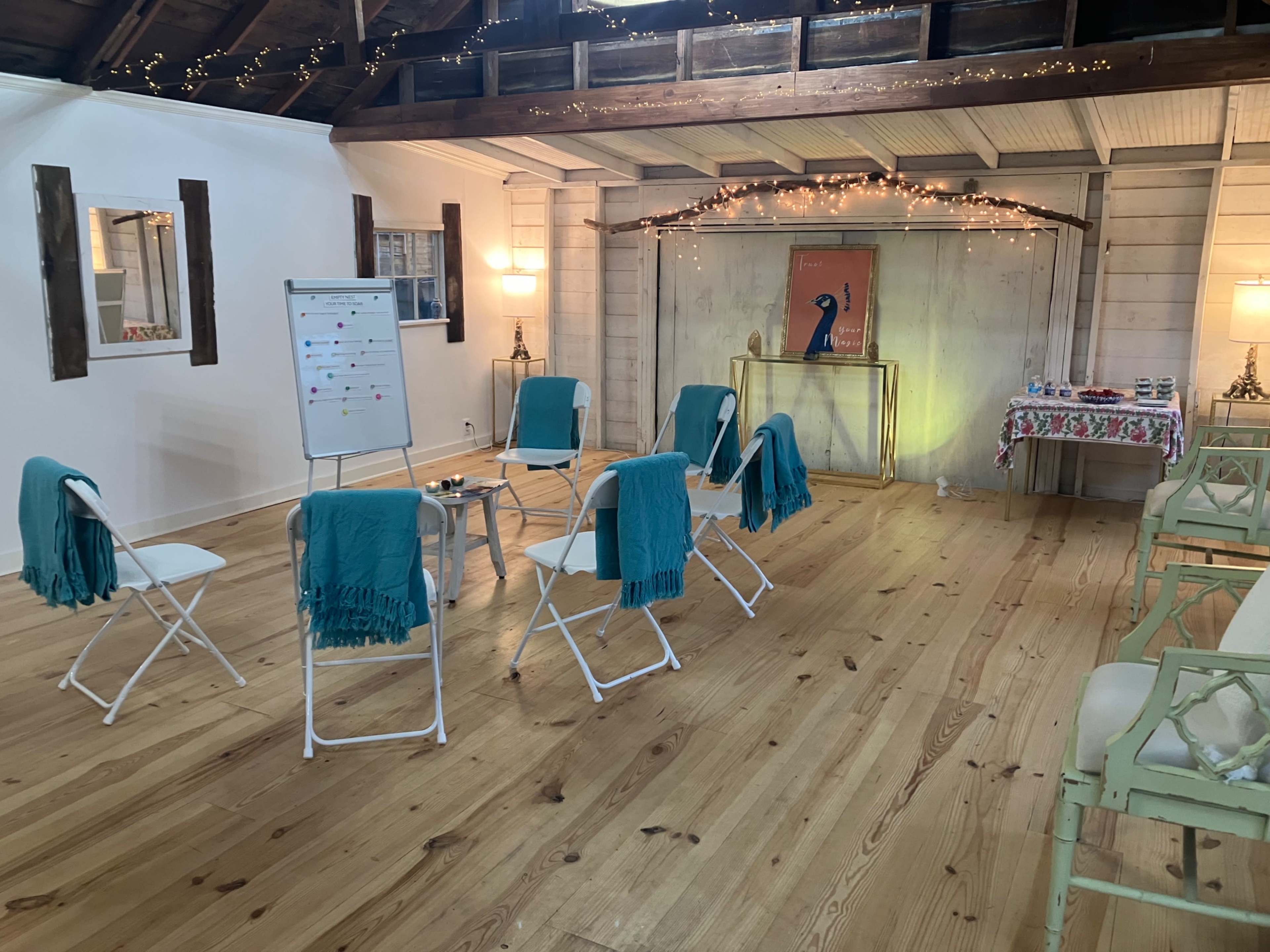 The image shows a well-lit room with wooden floors, featuring several folding chairs arranged in a circle, each with a teal throw draped over it, and a small table in the center.