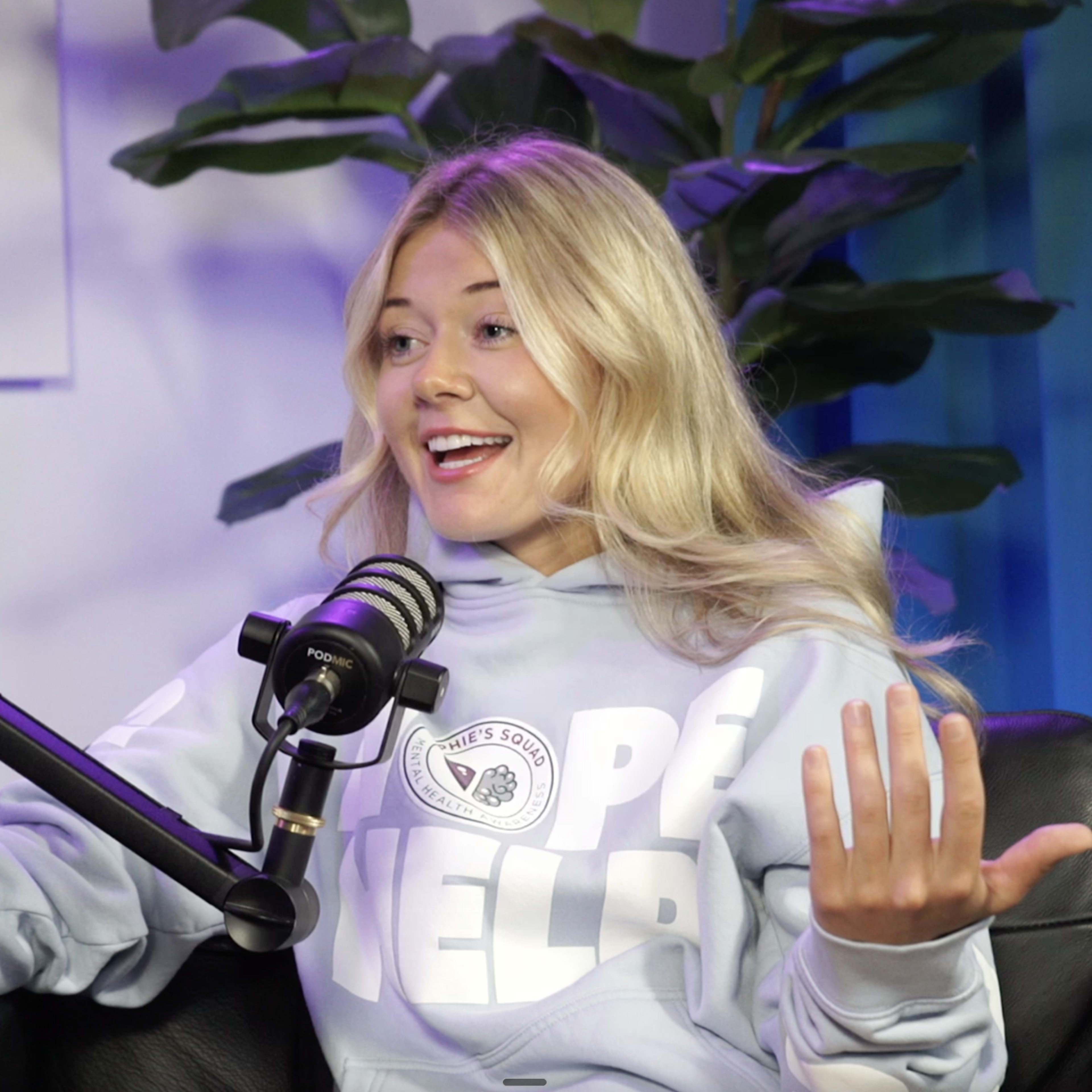 A young woman wearing a light blue hoodie is speaking animatedly in front of a microphone, surrounded by indoor plants.