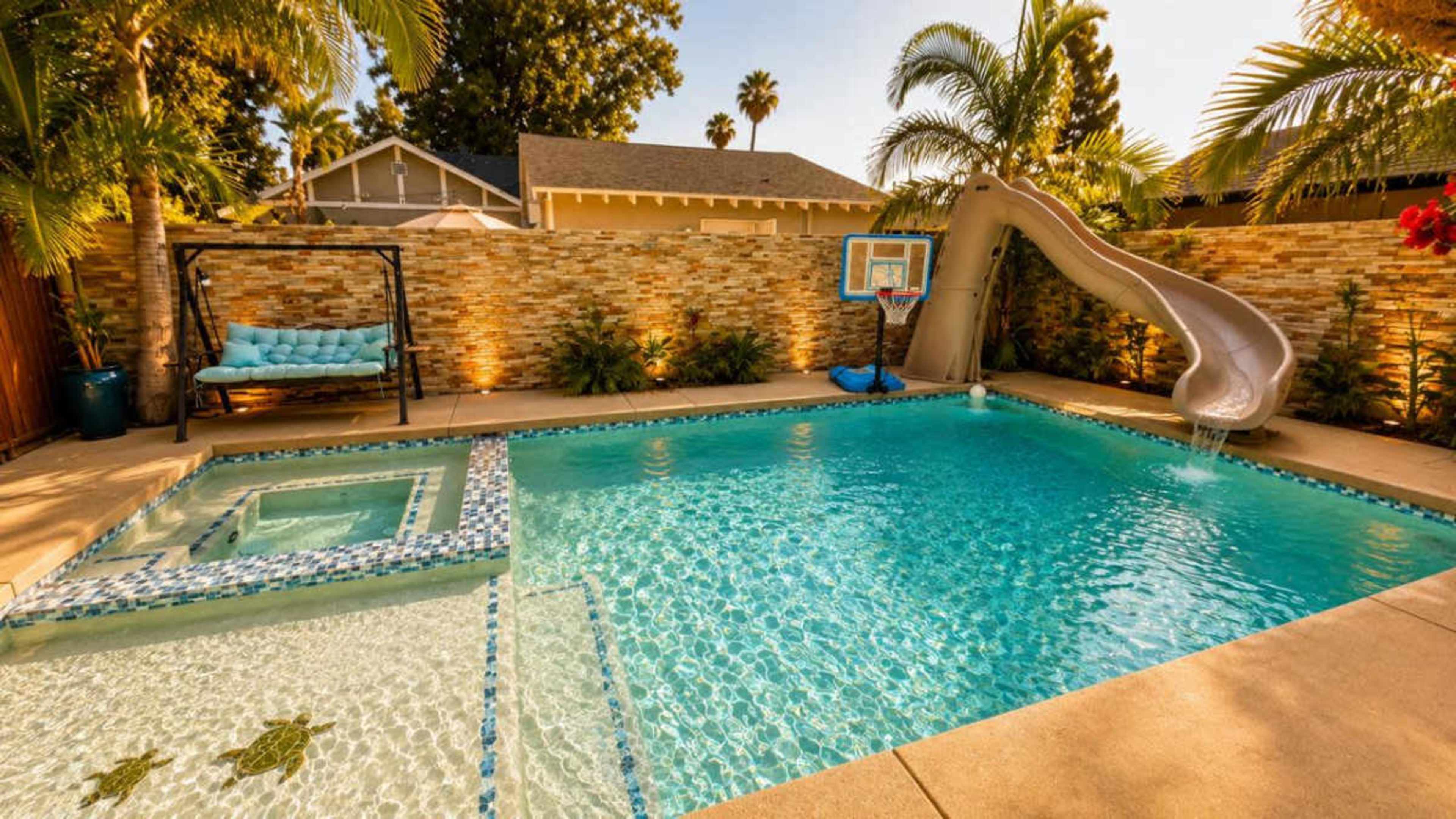 The image shows a backyard swimming pool with a water slide and a built-in spa, surrounded by palm trees and a stone wall.