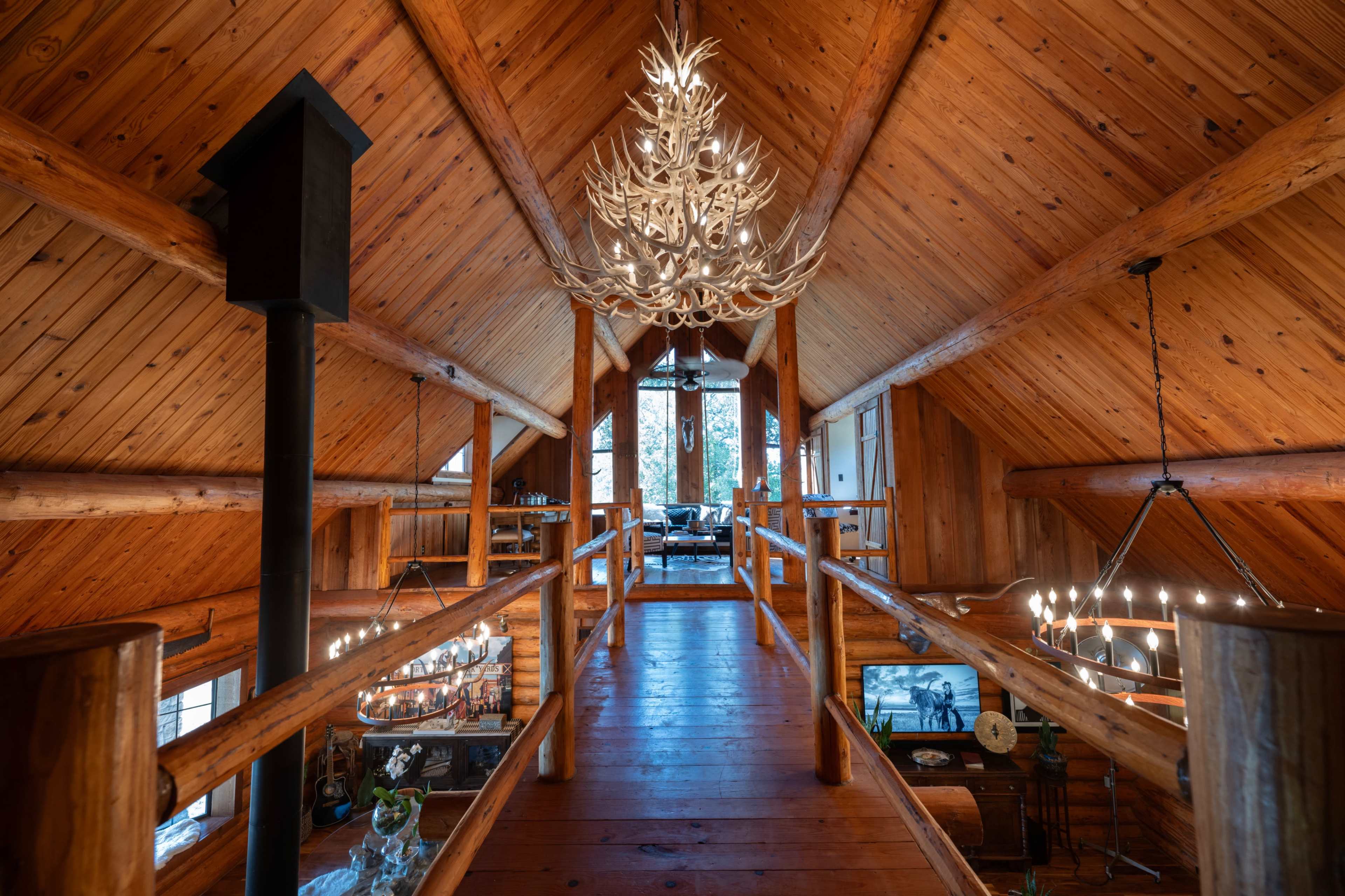 A wooden loft with a high ceiling features a large antler chandelier and multiple windows overlooking a living area below.
