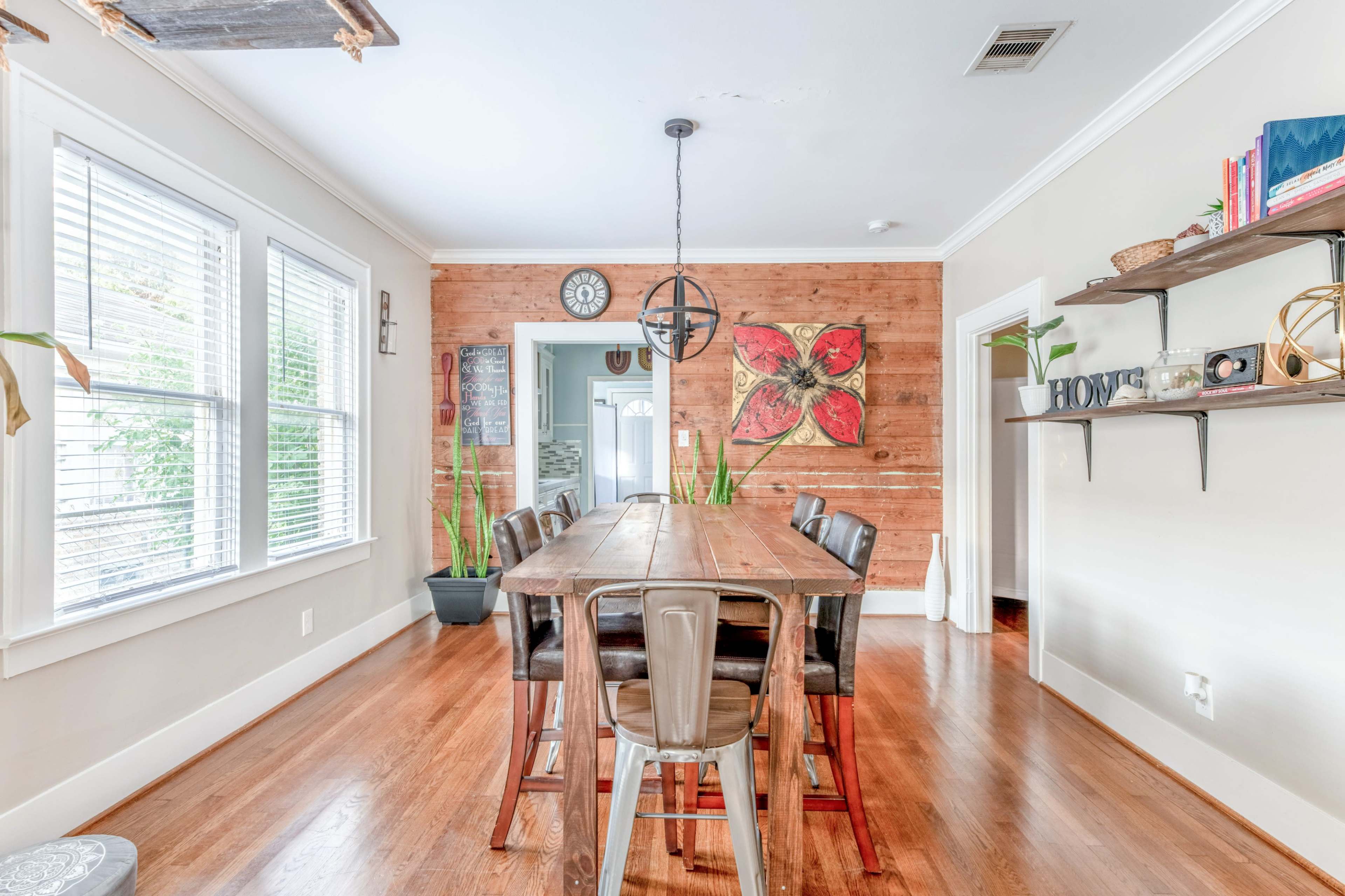 A wooden dining table with metal chairs is set against a backdrop of a light-colored wall and wood paneling, with plants and decor visible throughout the room.