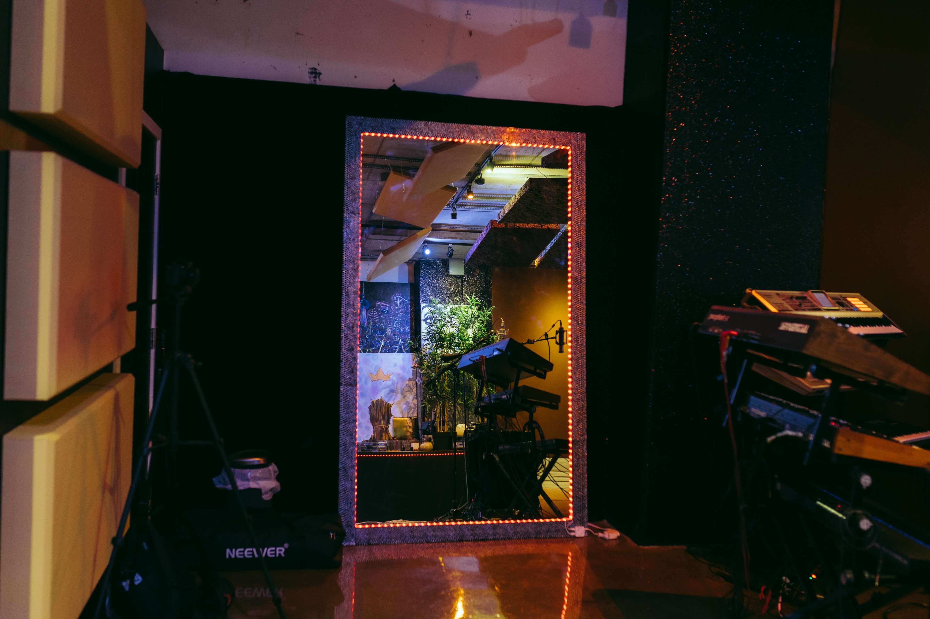 A large mirror framed with lights reflects a room containing musical instruments and plants, set against a backdrop of dark walls.