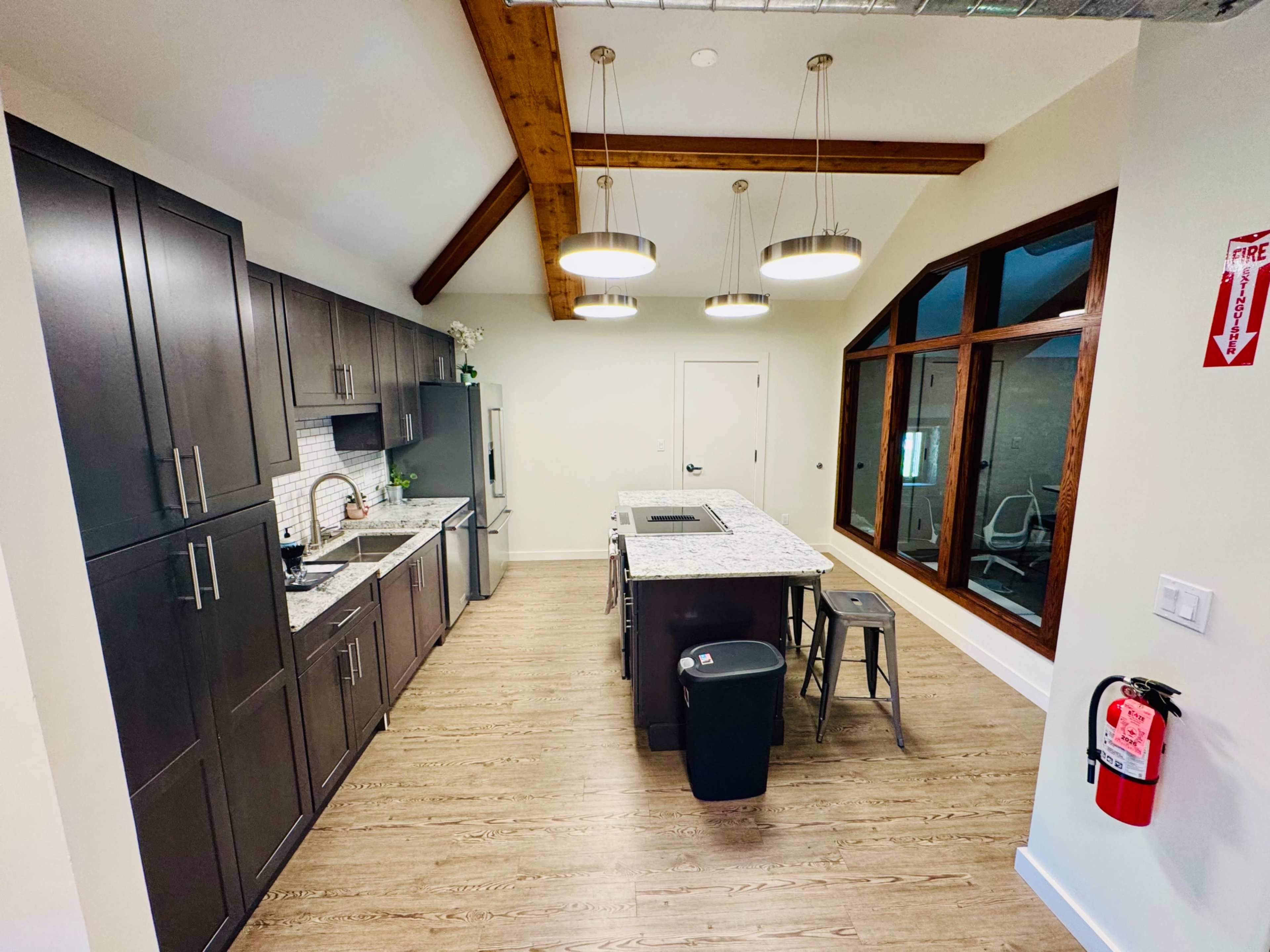 A modern kitchen features dark cabinets, a granite island, and large windows with wooden beams overhead.
