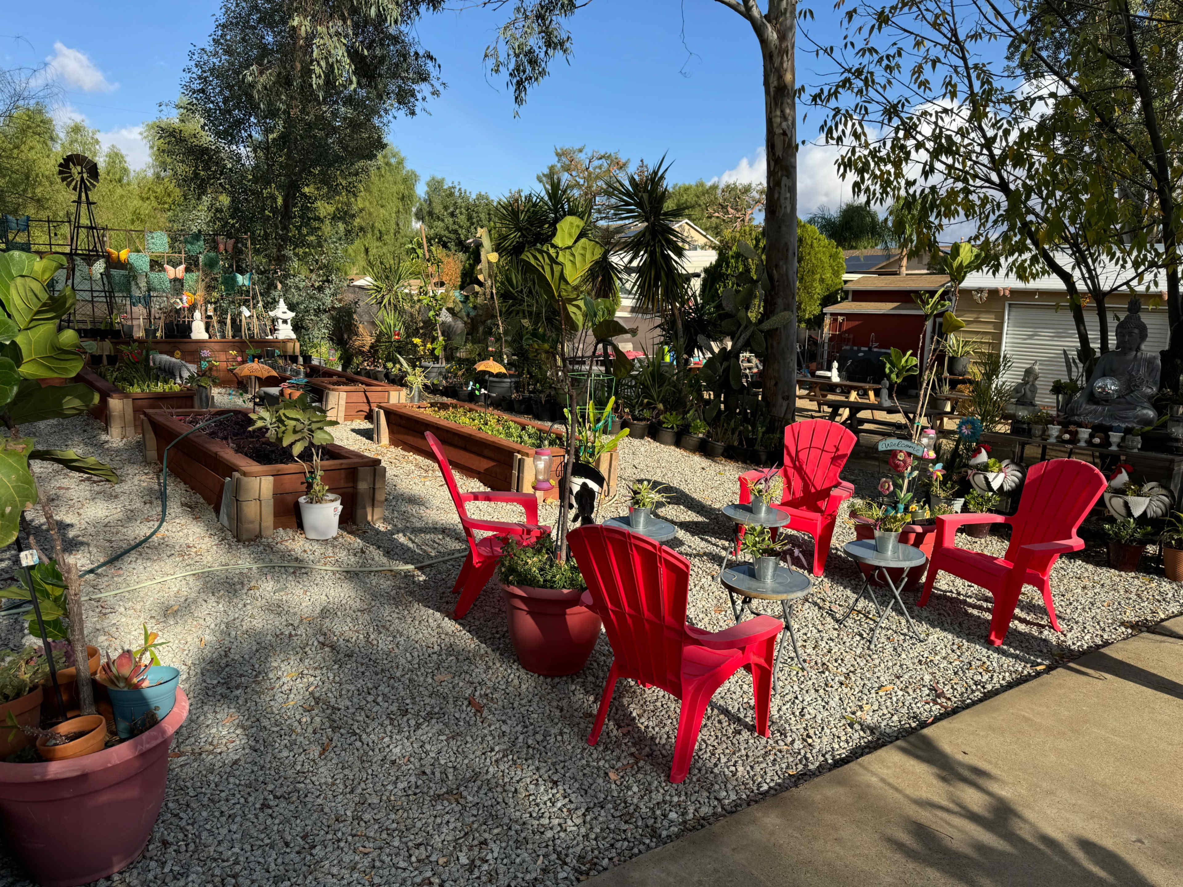 A garden area features red outdoor chairs and tables surrounded by various plants and decorative items on a gravel surface.