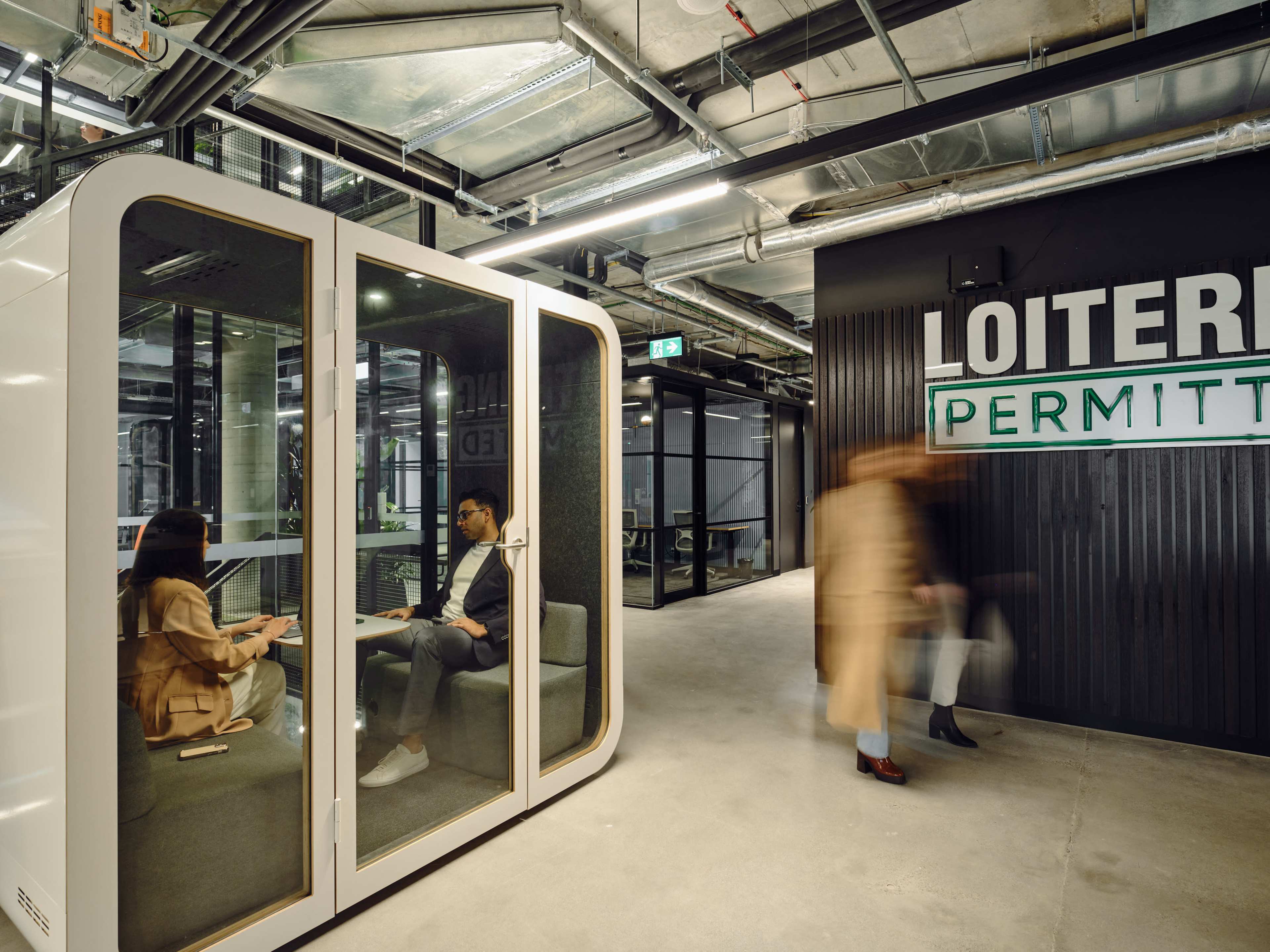 The image shows a modern office space with a white meeting booth on the left, where two people are engaged in a discussion, and a sign on the right that reads "LOITERING PERMITTED."