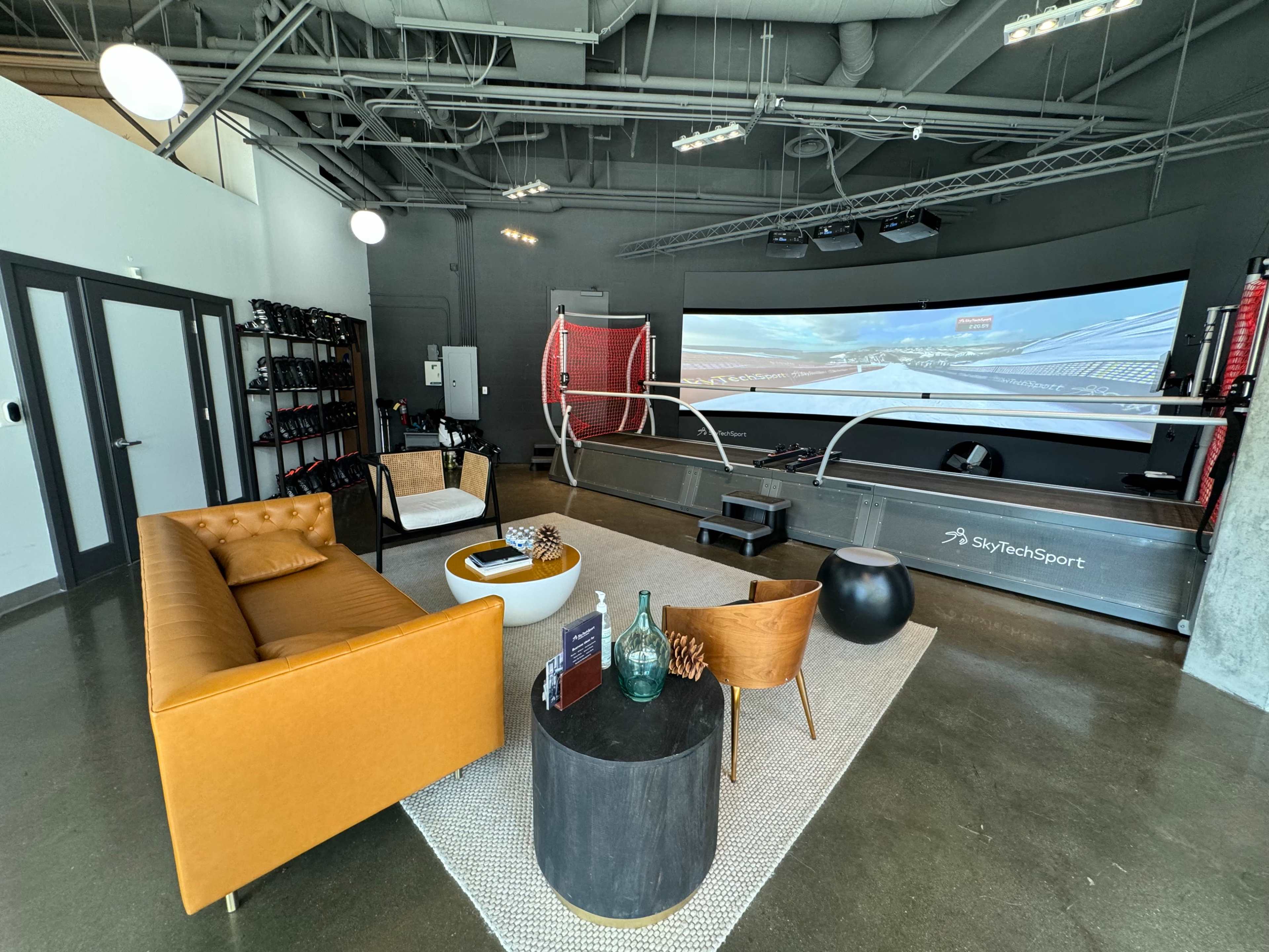 The image shows a modern lounge area with a brown sofa, a round coffee table, and a large screen displaying a racing simulation setup.