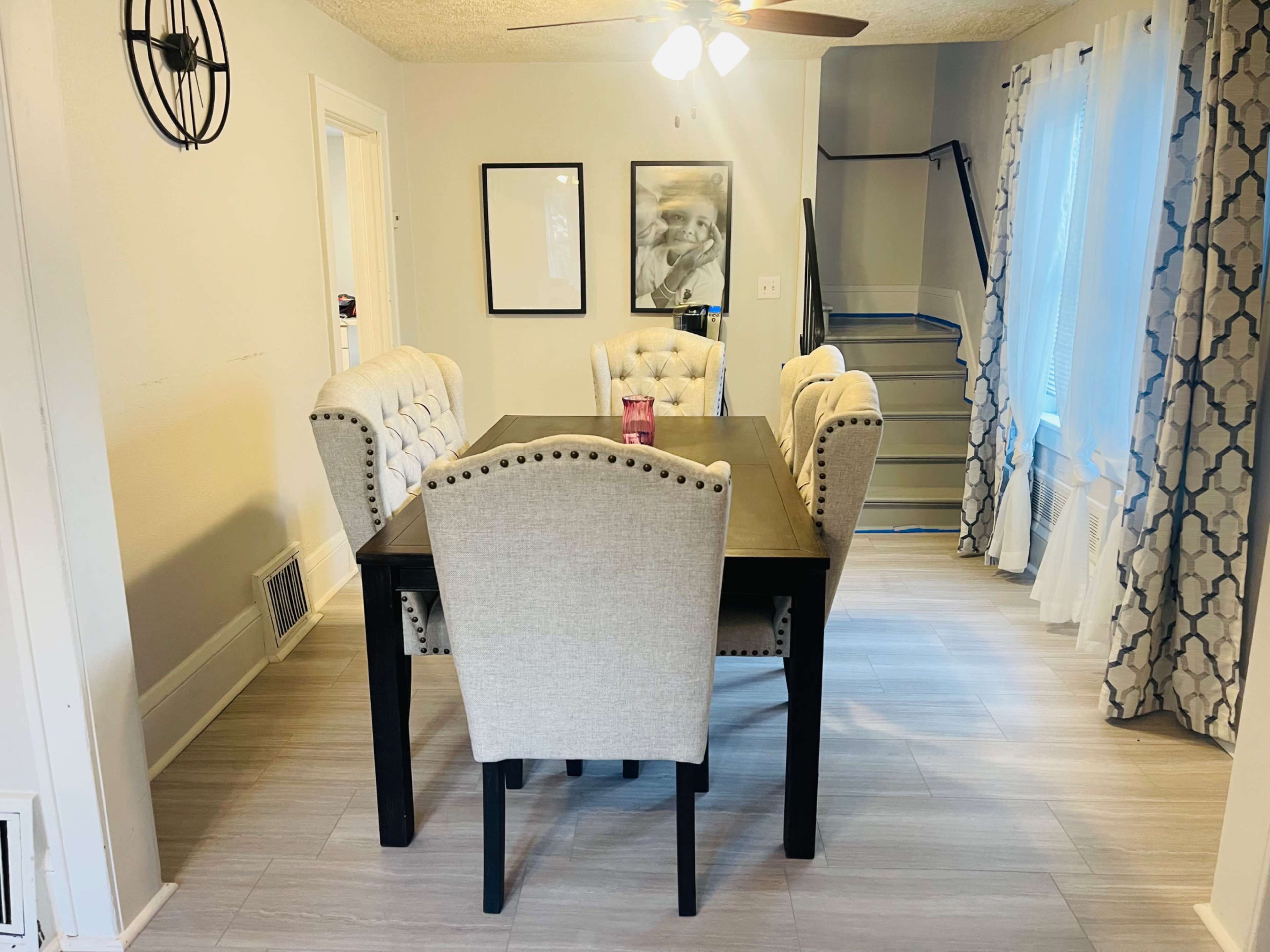A dining room with a rectangular table surrounded by four upholstered chairs, a ceiling fan above, and a staircase visible in the background.