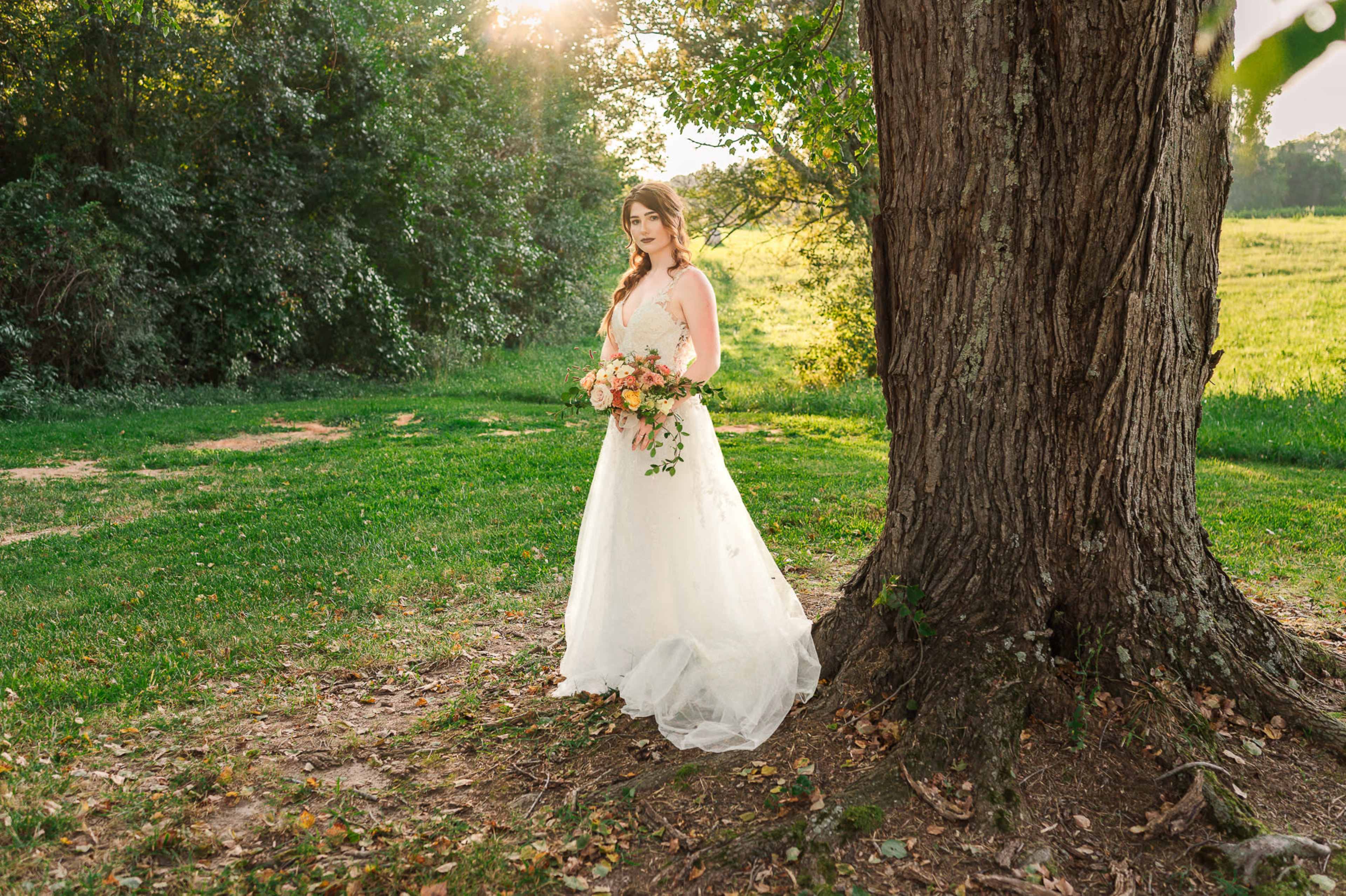 A bride stands by a large tree in a grassy field, holding a bouquet of flowers.