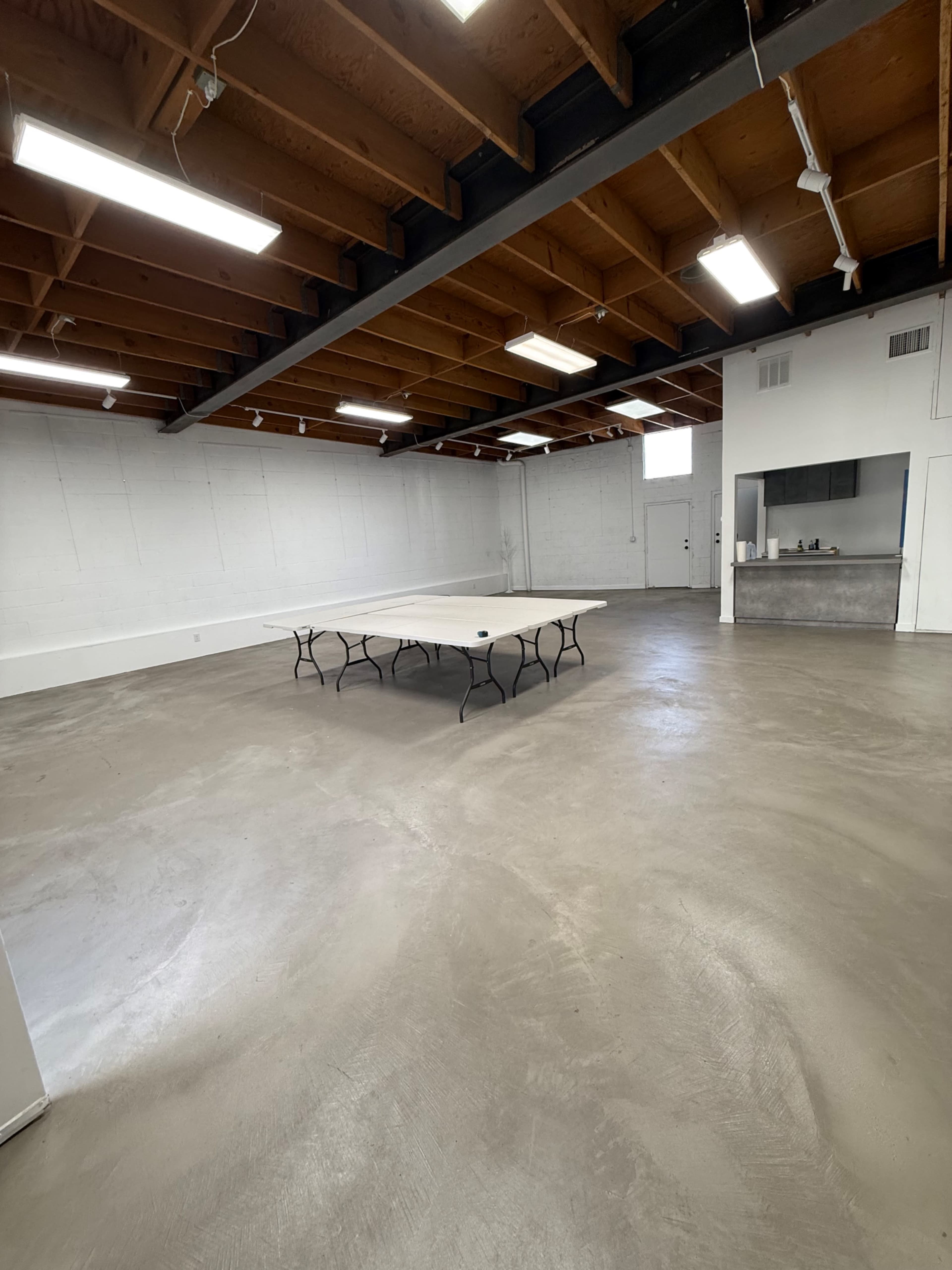 The image shows a spacious, empty room with a table in the center and a kitchenette area in the background.