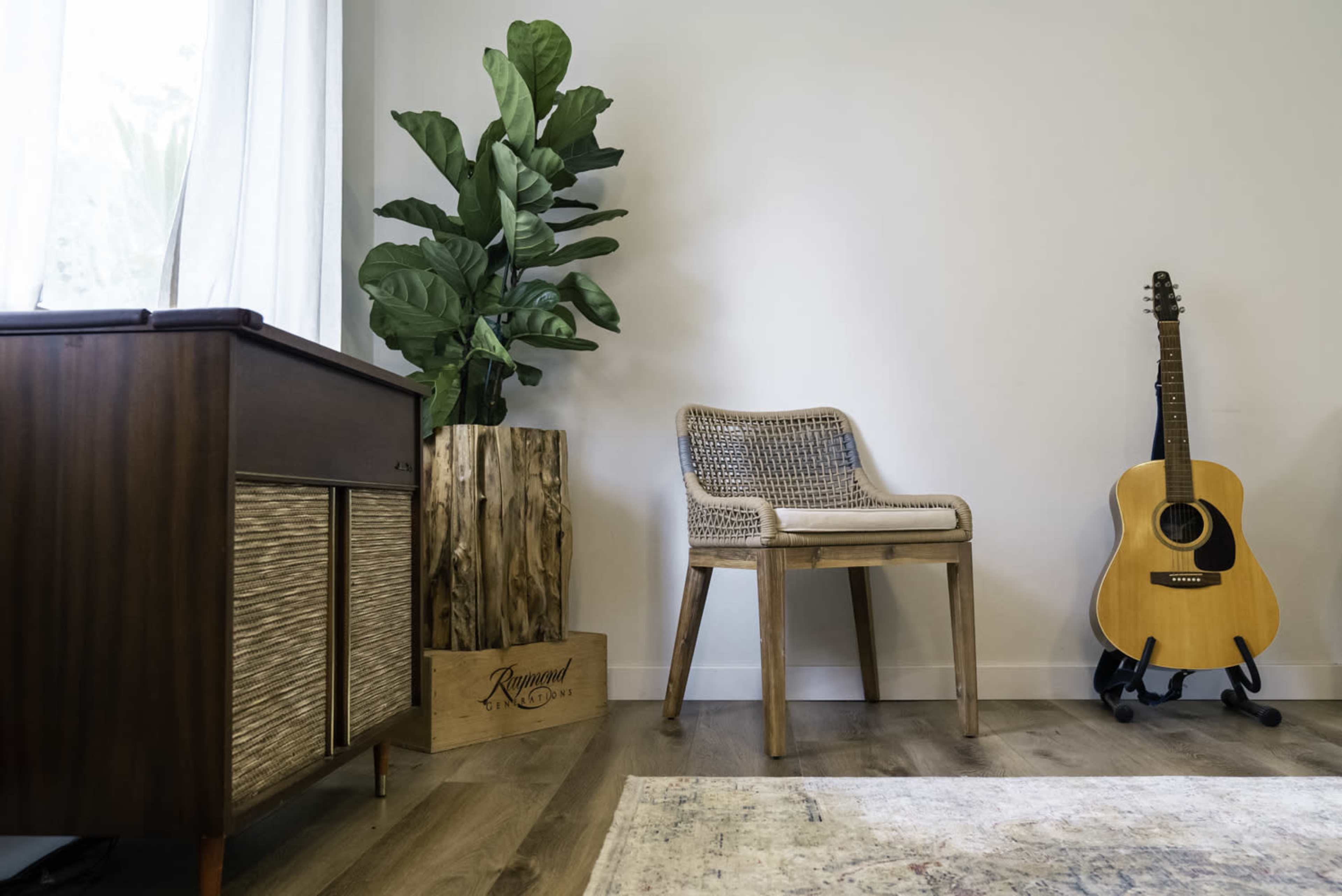 A wooden chair and an acoustic guitar stand beside a tall potted plant in a minimalist room with light-colored walls.