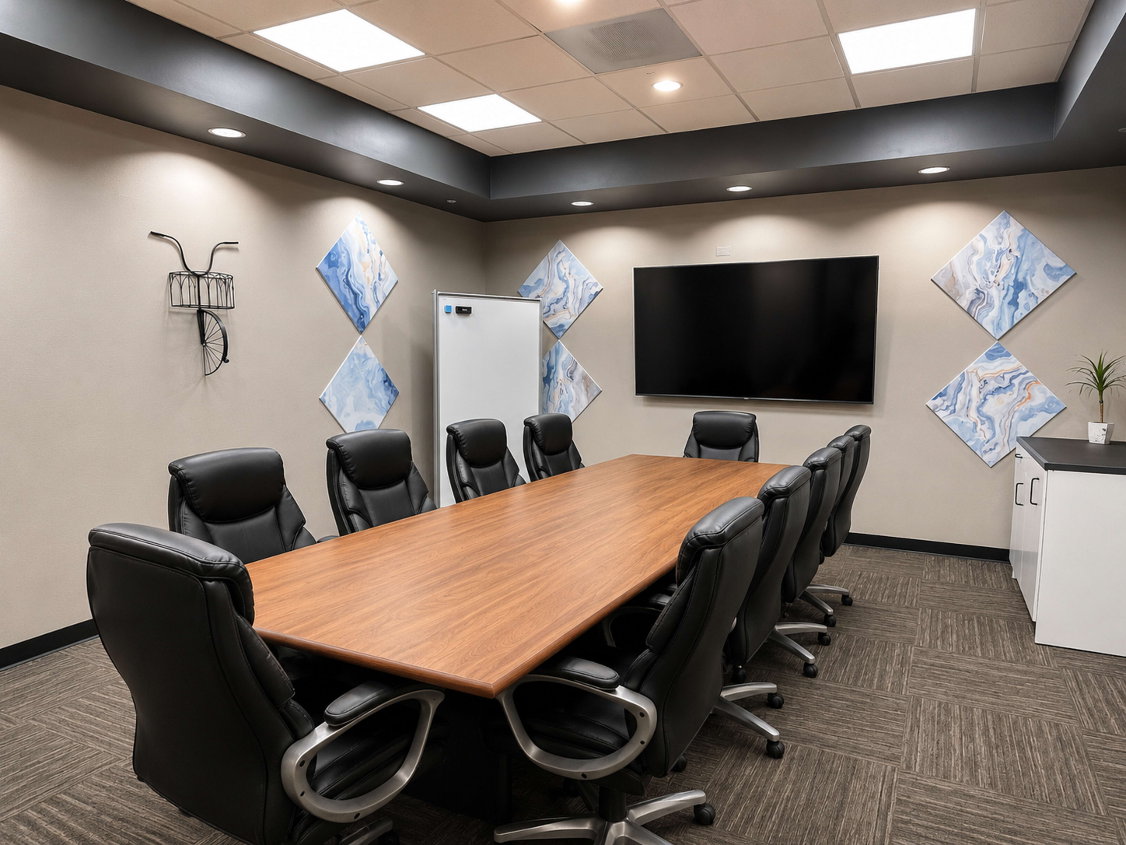 A modern conference room features a large wooden table surrounded by black leather chairs, a mounted television, and abstract artwork on the walls.
