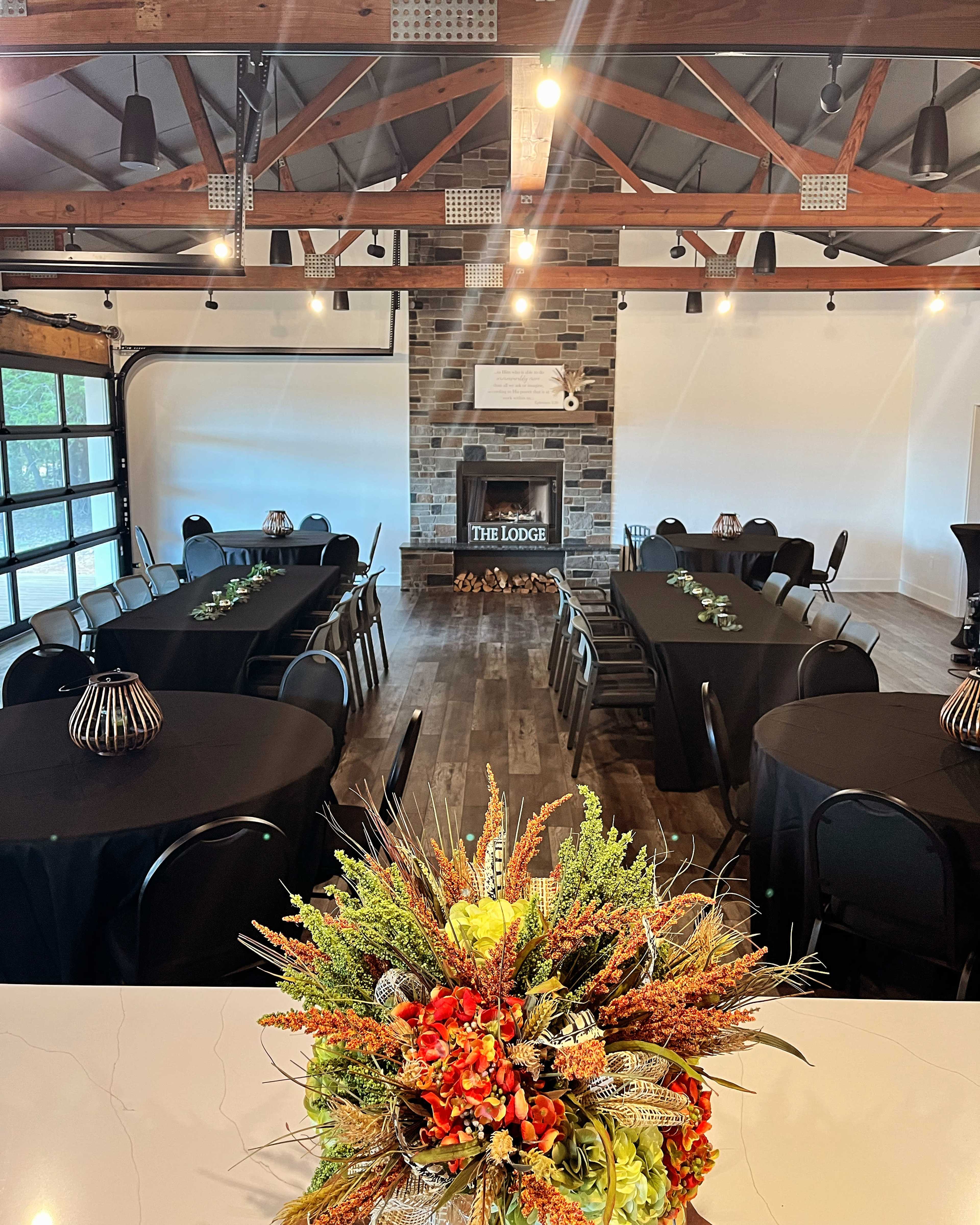 The image shows an indoor event space with black tables arranged in a U-shape, a stone fireplace at the back, and a decorative floral arrangement in the foreground.