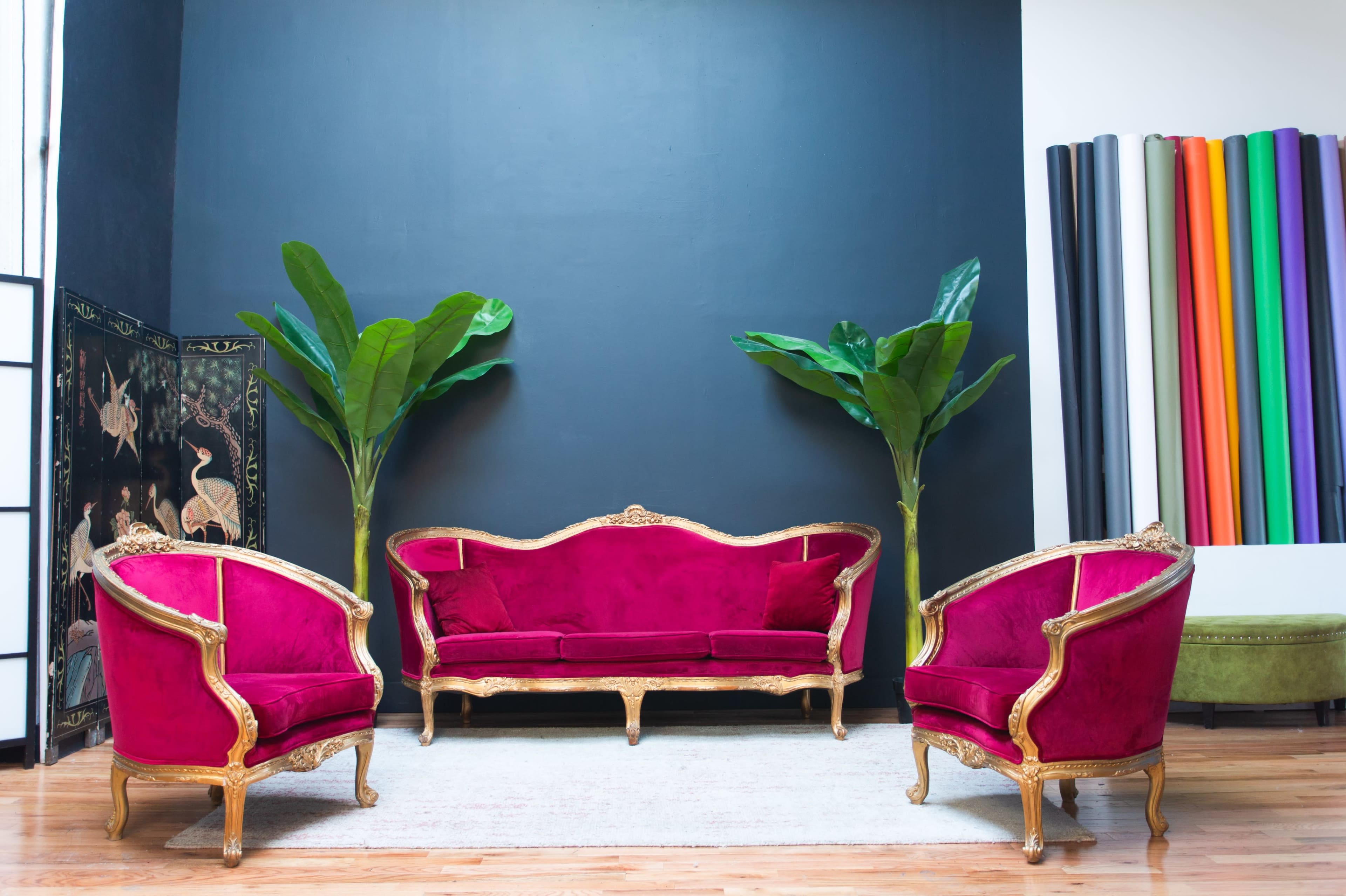 The image shows a vibrant seating arrangement with a pink velvet sofa and two matching chairs, framed by large green plants and a colorful vertical display on the wall.
