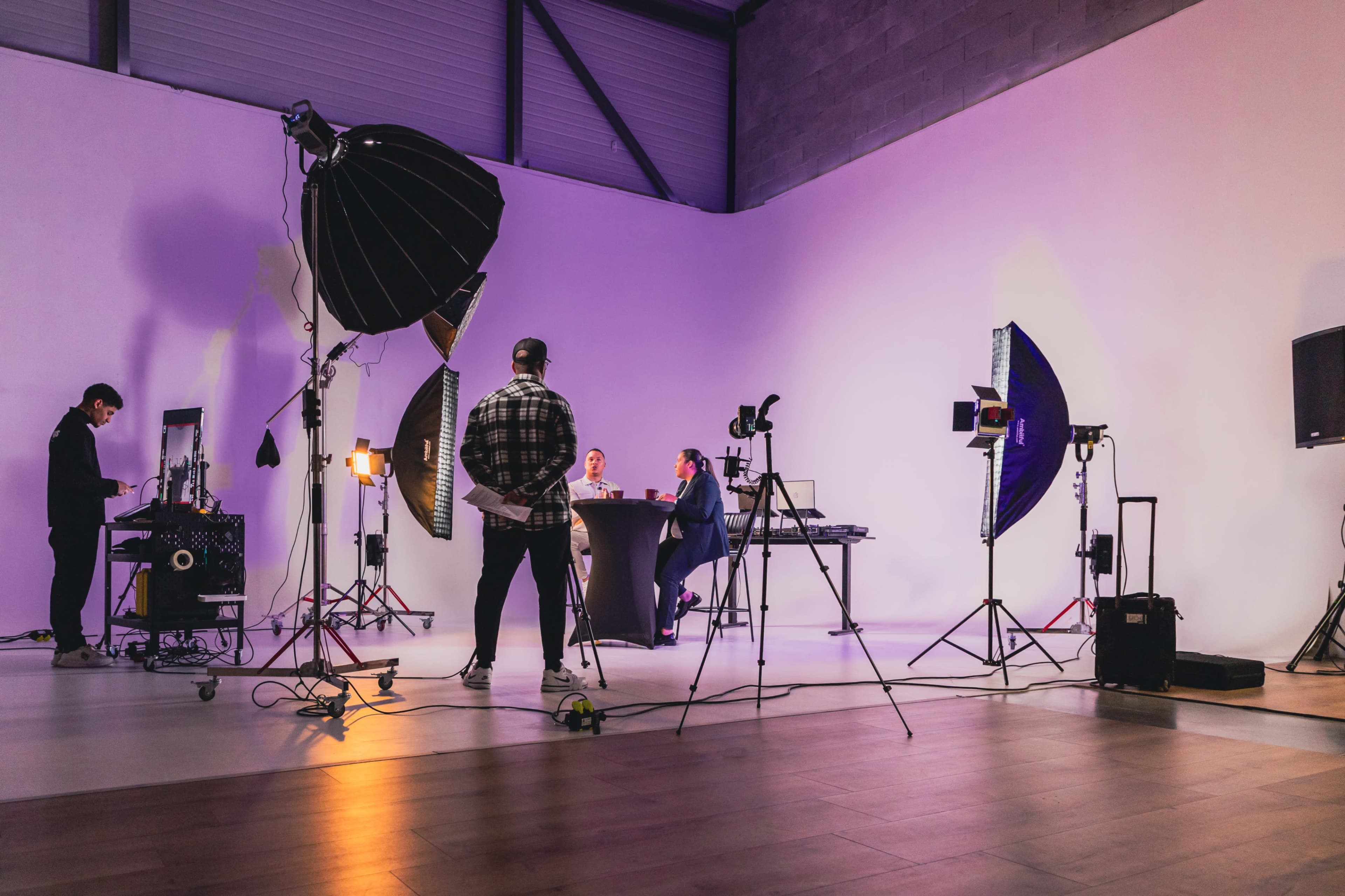 A group of people is seated at a table in a studio with professional lighting and cameras set up for a shoot.