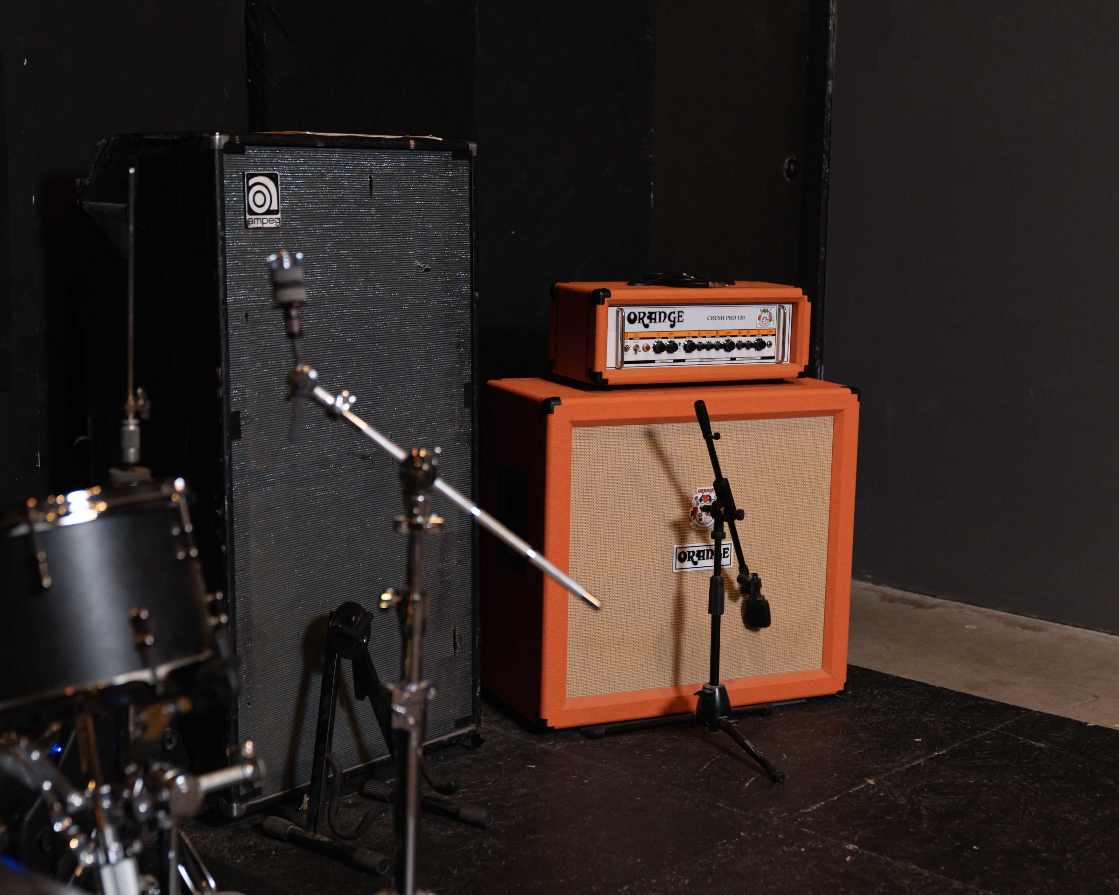 The image shows a music practice space with a black amplifier cabinet, an orange guitar amplifier head, and a drum kit setup nearby.