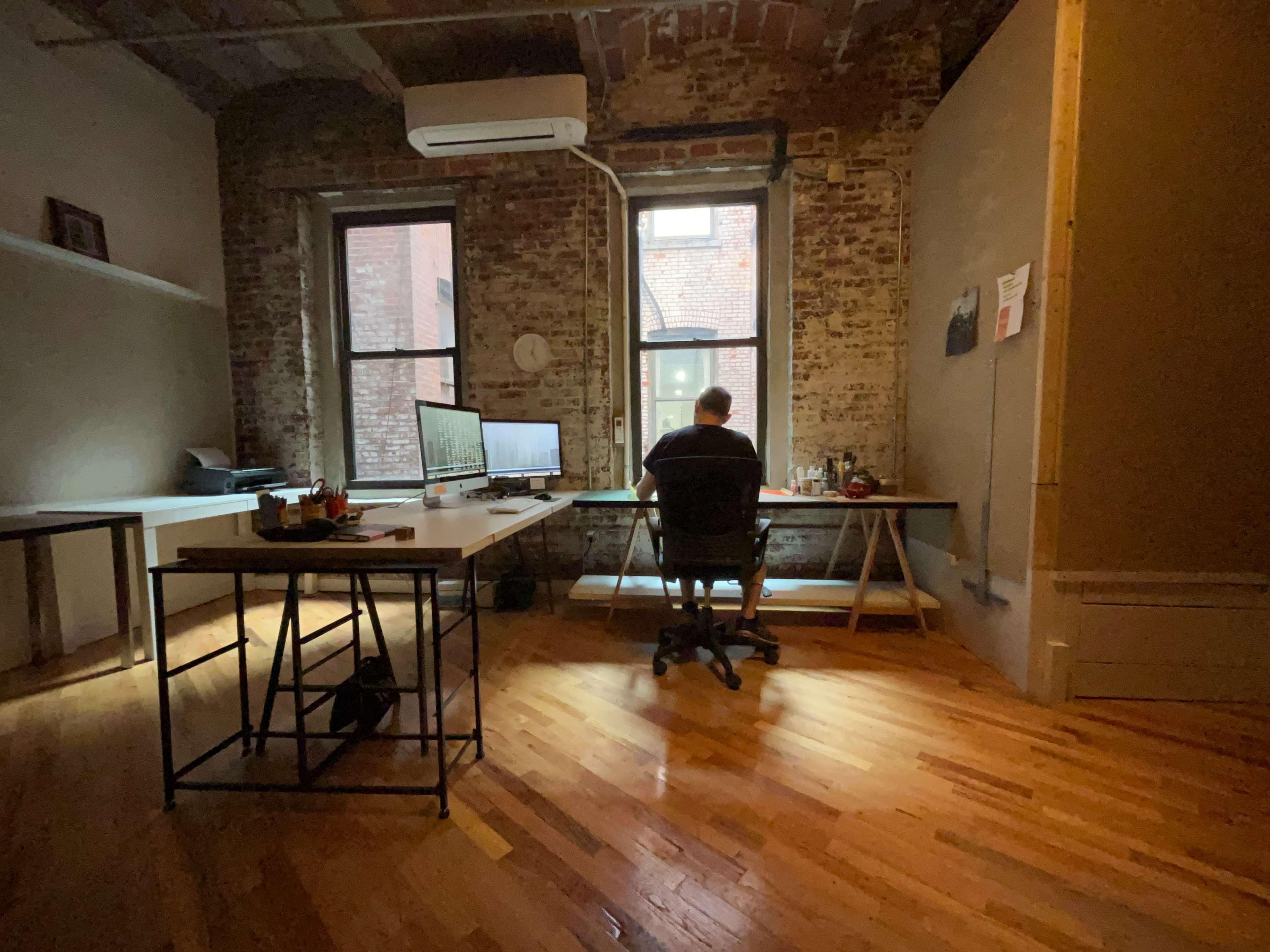 A person is seated at a desk in a well-lit room with exposed brick walls and large windows.