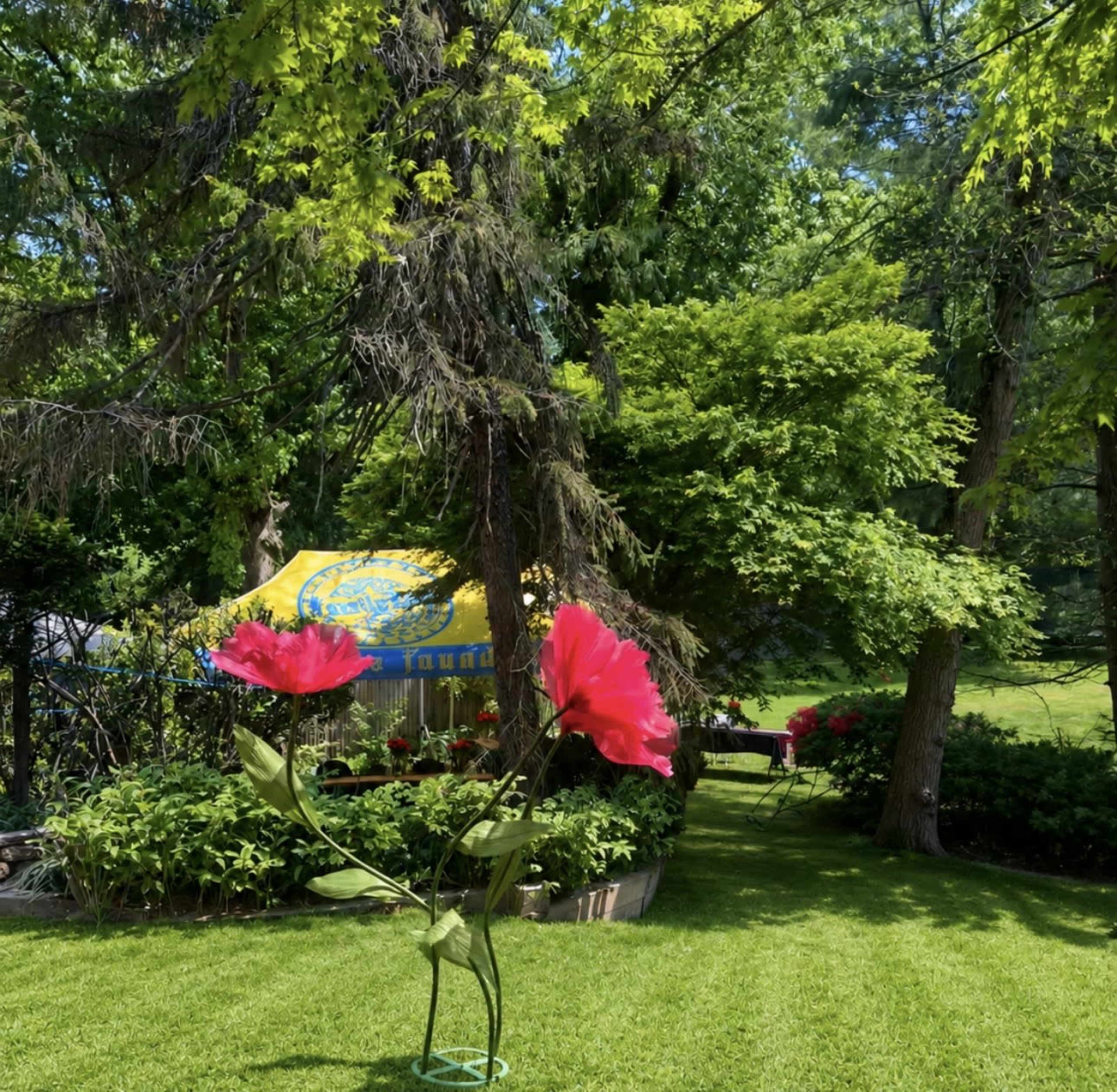 A large pink flower sculpture stands amidst a neatly manicured lawn and lush greenery, with a colorful tent in the background.