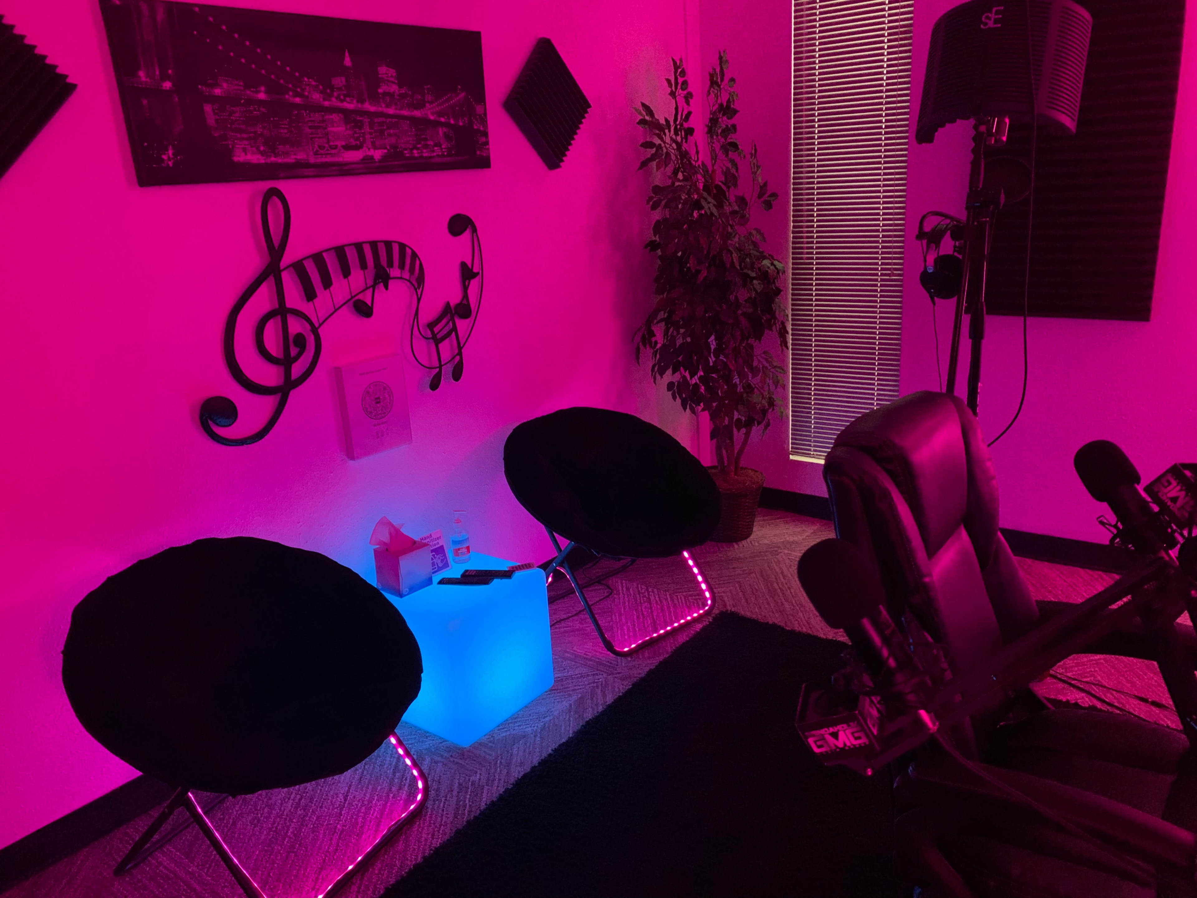 A room with pink lighting, featuring two black chairs, a blue illuminated table, and wall decor of musical notes and a cityscape.