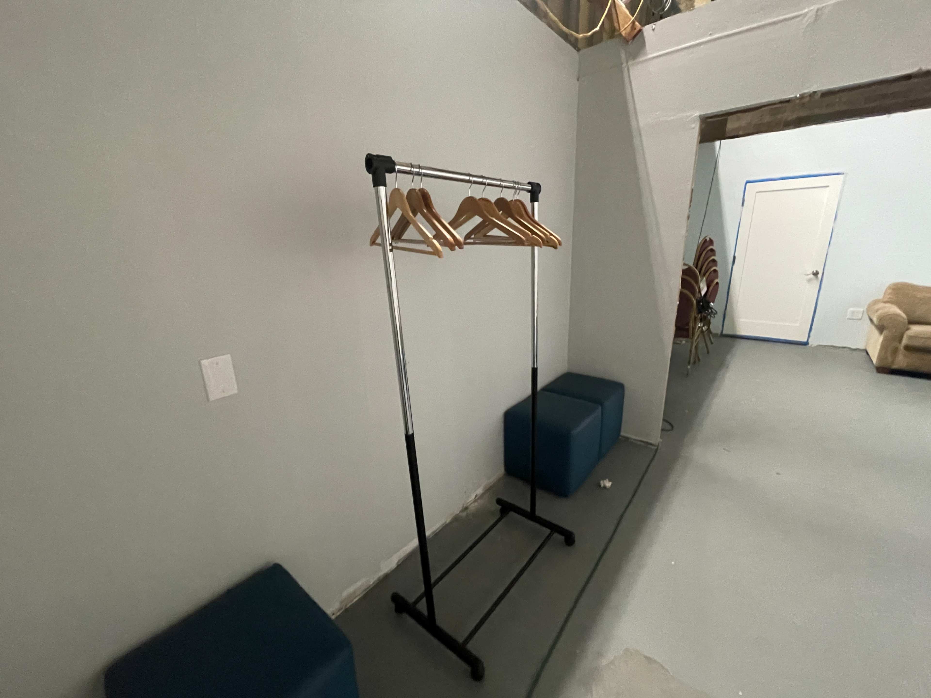 A clothing rack with wooden hangers stands against a gray wall in a sparsely furnished room.
