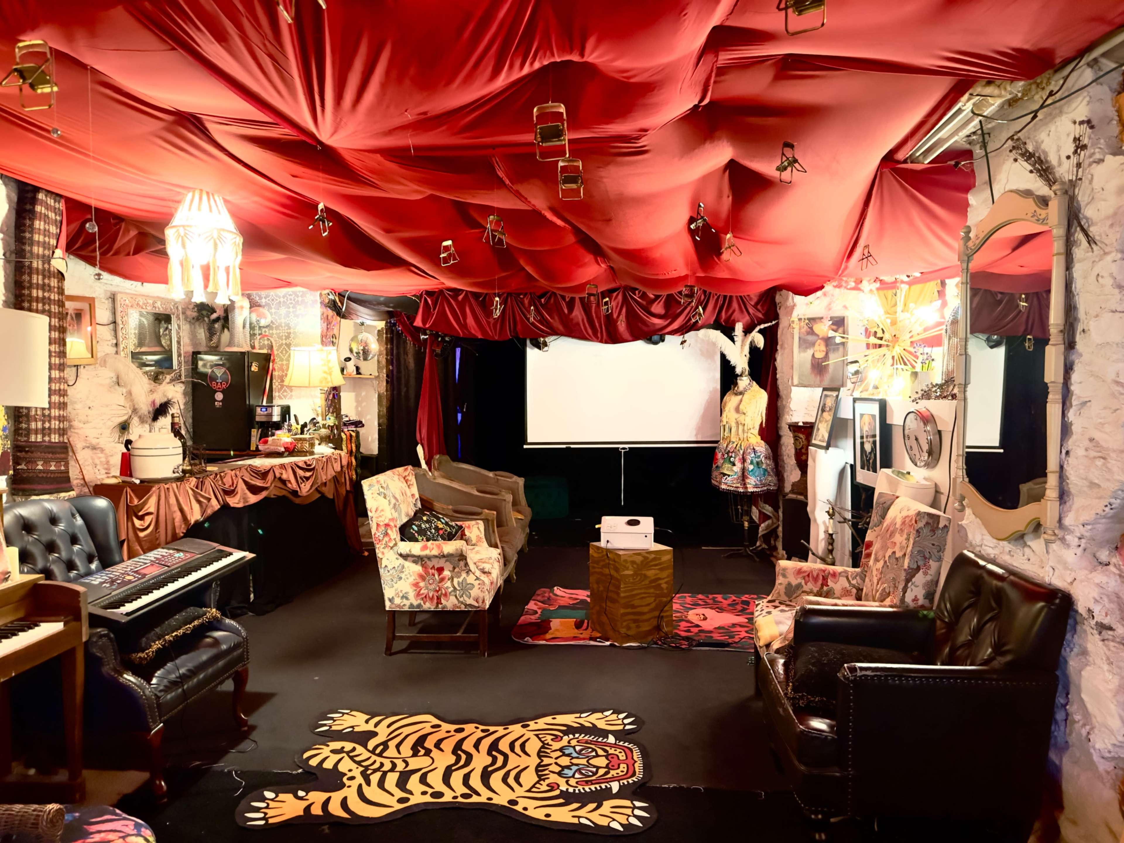 The room features eclectic furnishings with a red fabric ceiling, patterned rugs, and a projection screen at the front.