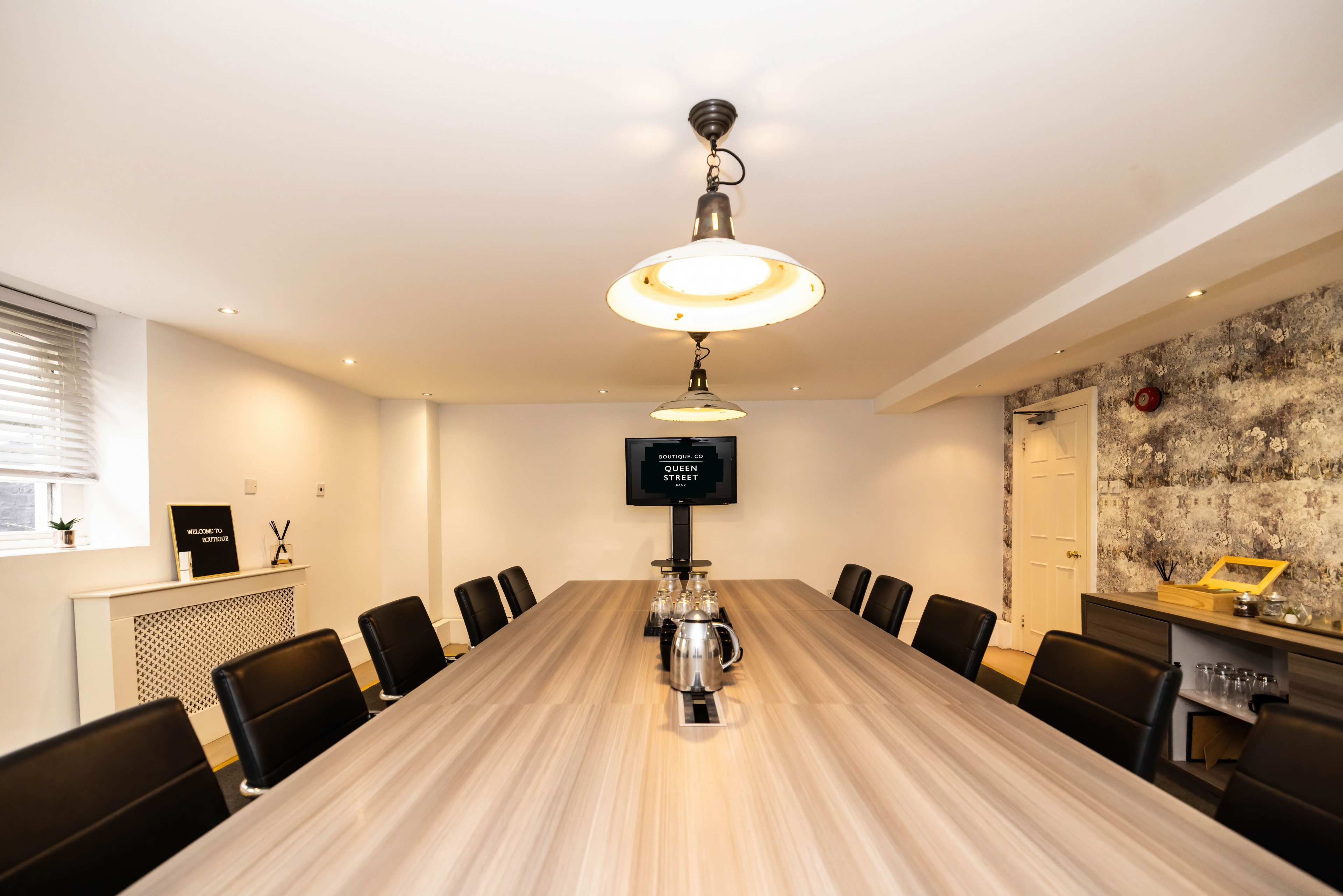 A modern conference room features a long wooden table surrounded by black leather chairs, with a screen mounted on the wall displaying a message.