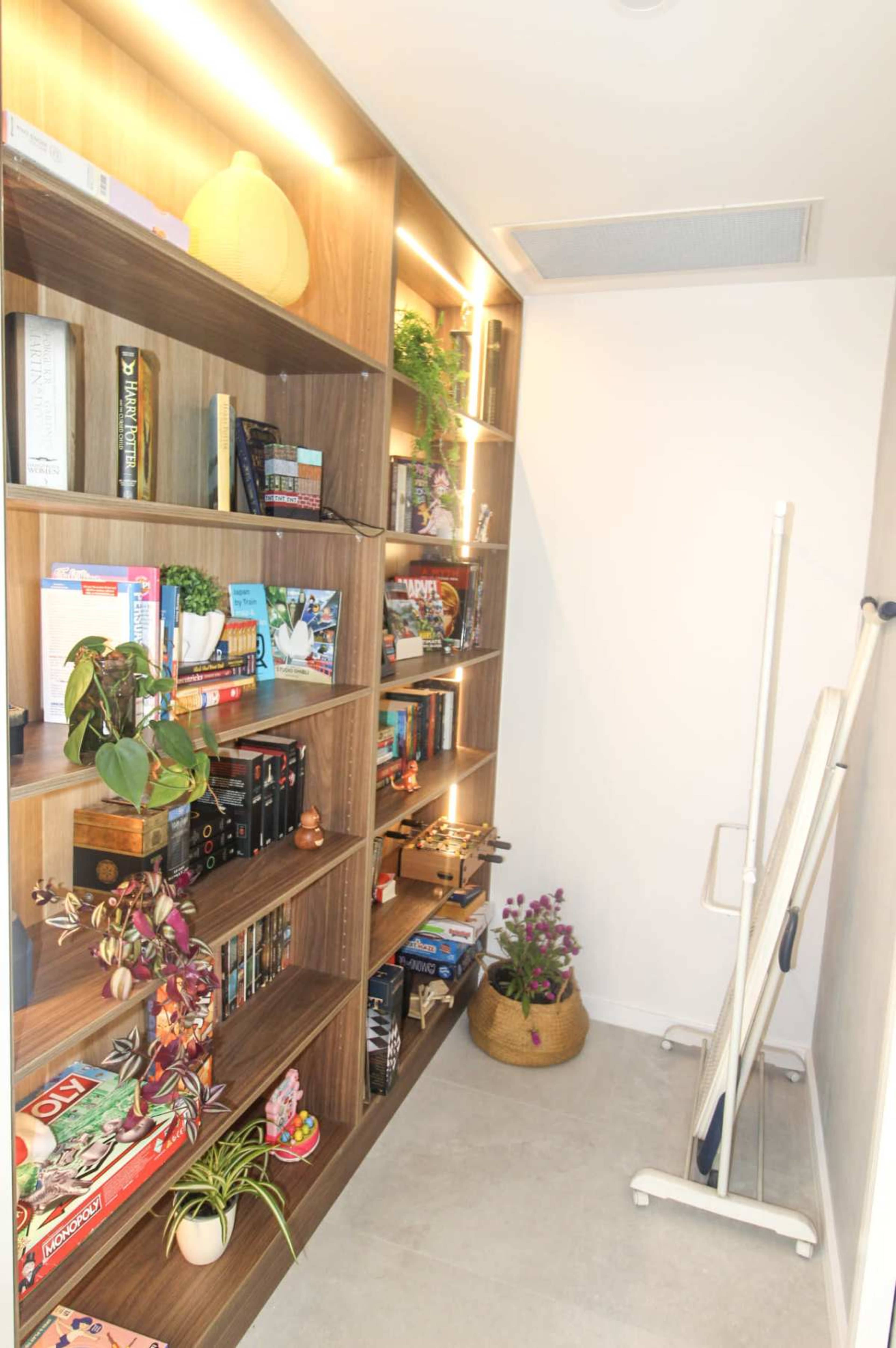 The image shows a small study nook with wooden bookshelves filled with books and decorative plants, along with a corner displaying a broom and a basket of flowers.