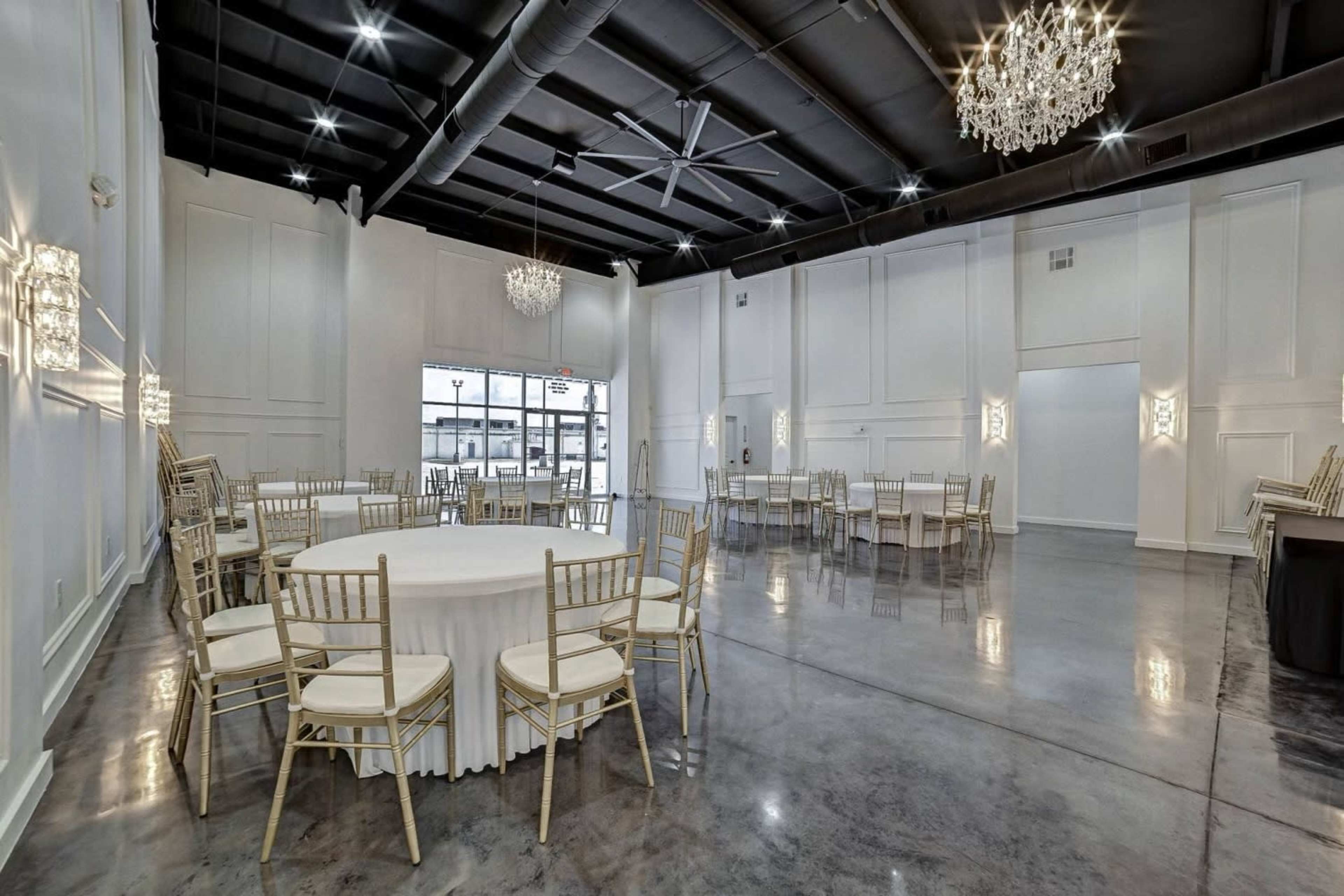 A spacious event hall features round tables with white tablecloths surrounded by gold chairs under elegant chandeliers.