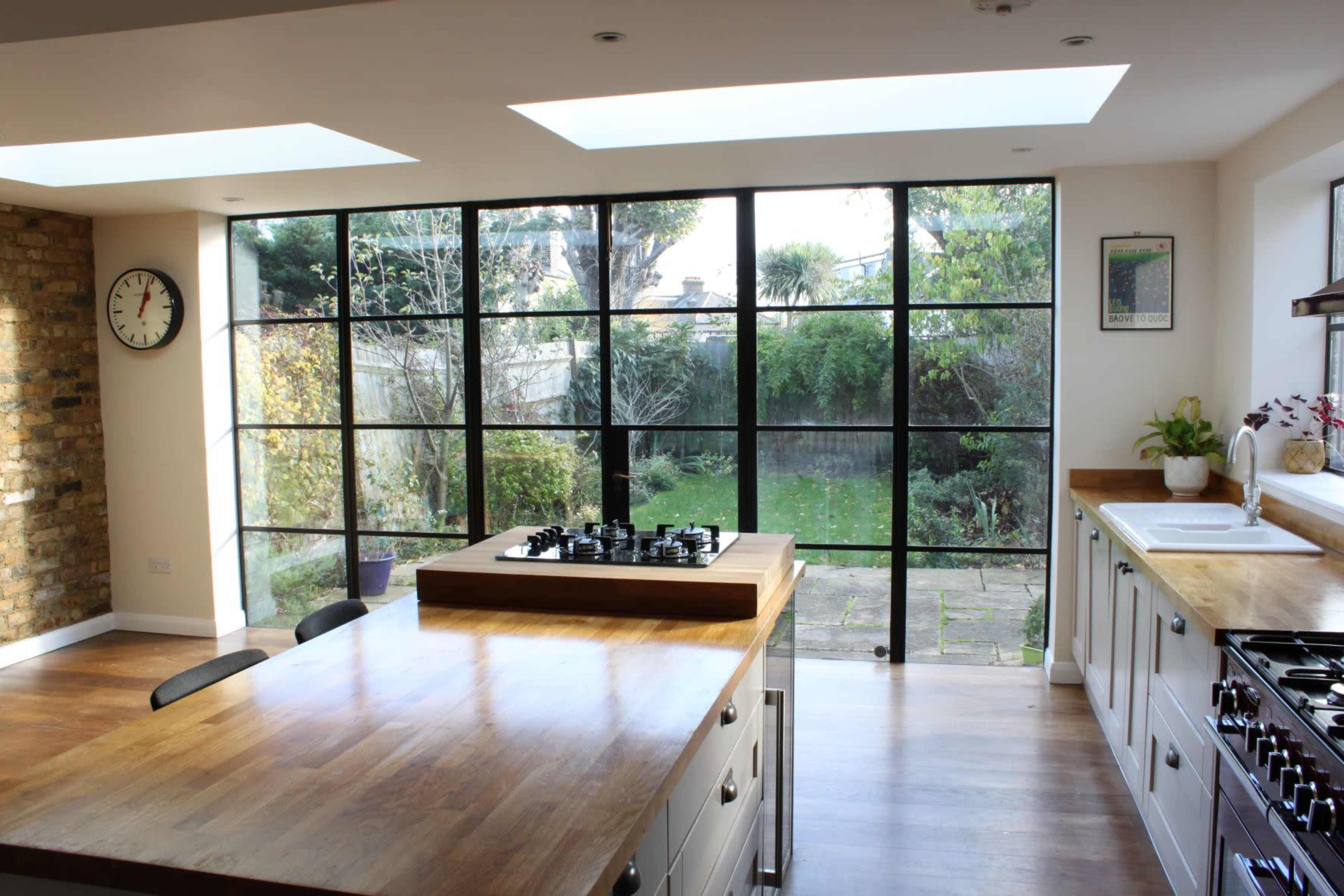 A modern kitchen with a central wooden island, large windows overlooking a garden, and a brick wall.