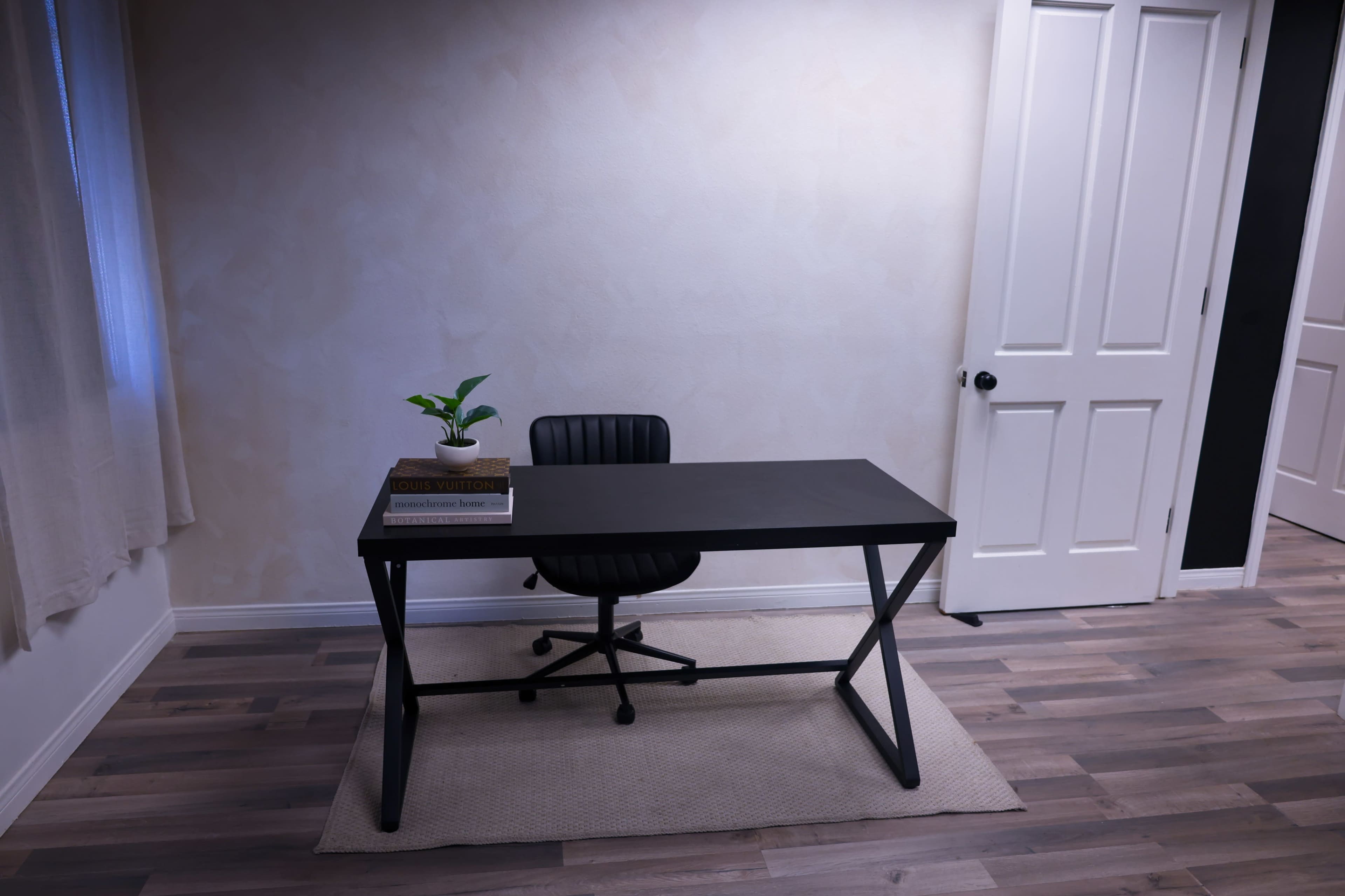 A black desk with a chair sits on a beige rug in a room with light-colored walls and a closed white door.